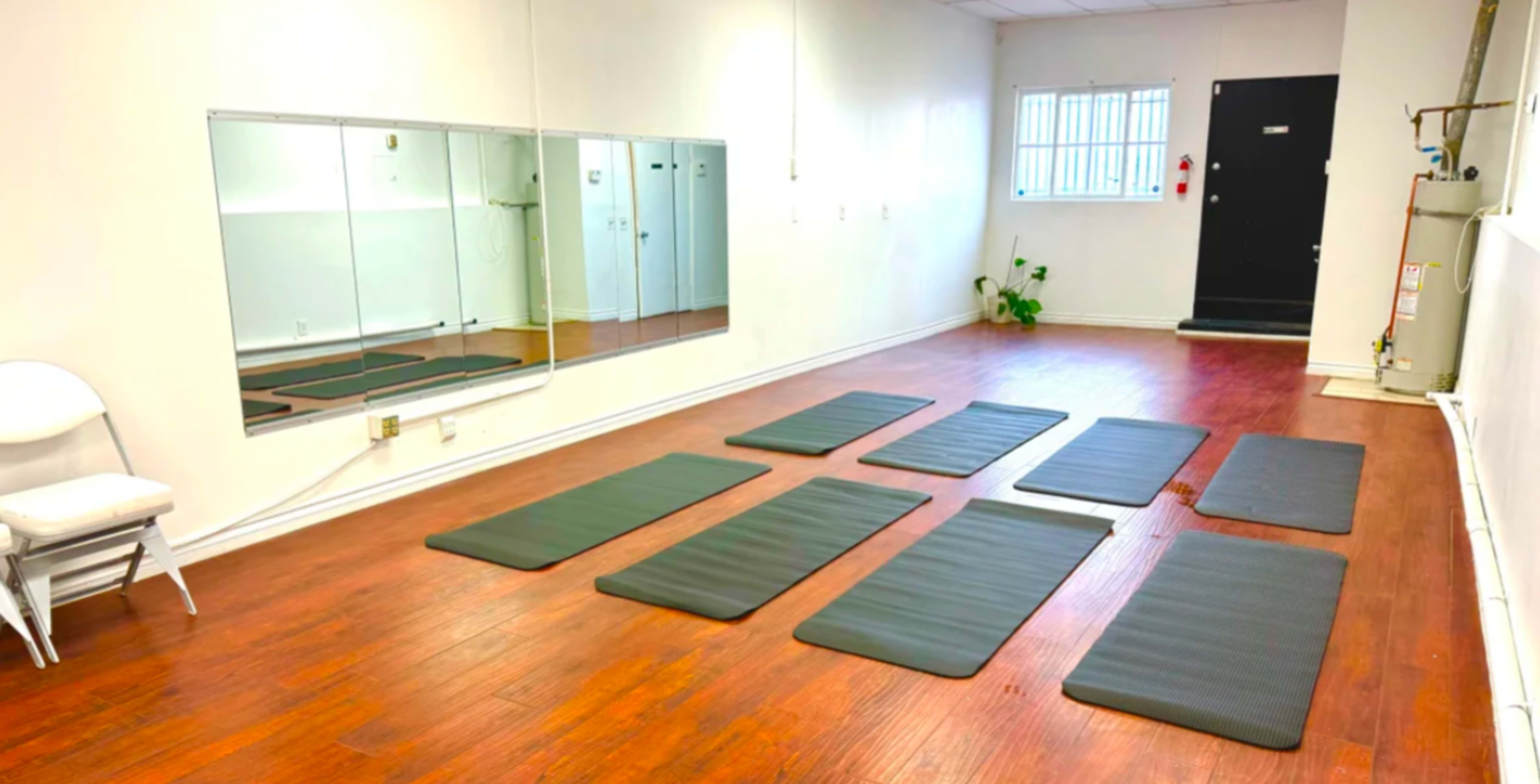 A spacious, well-lit exercise room with several black mats arranged on a wooden floor and a large mirror along one wall.