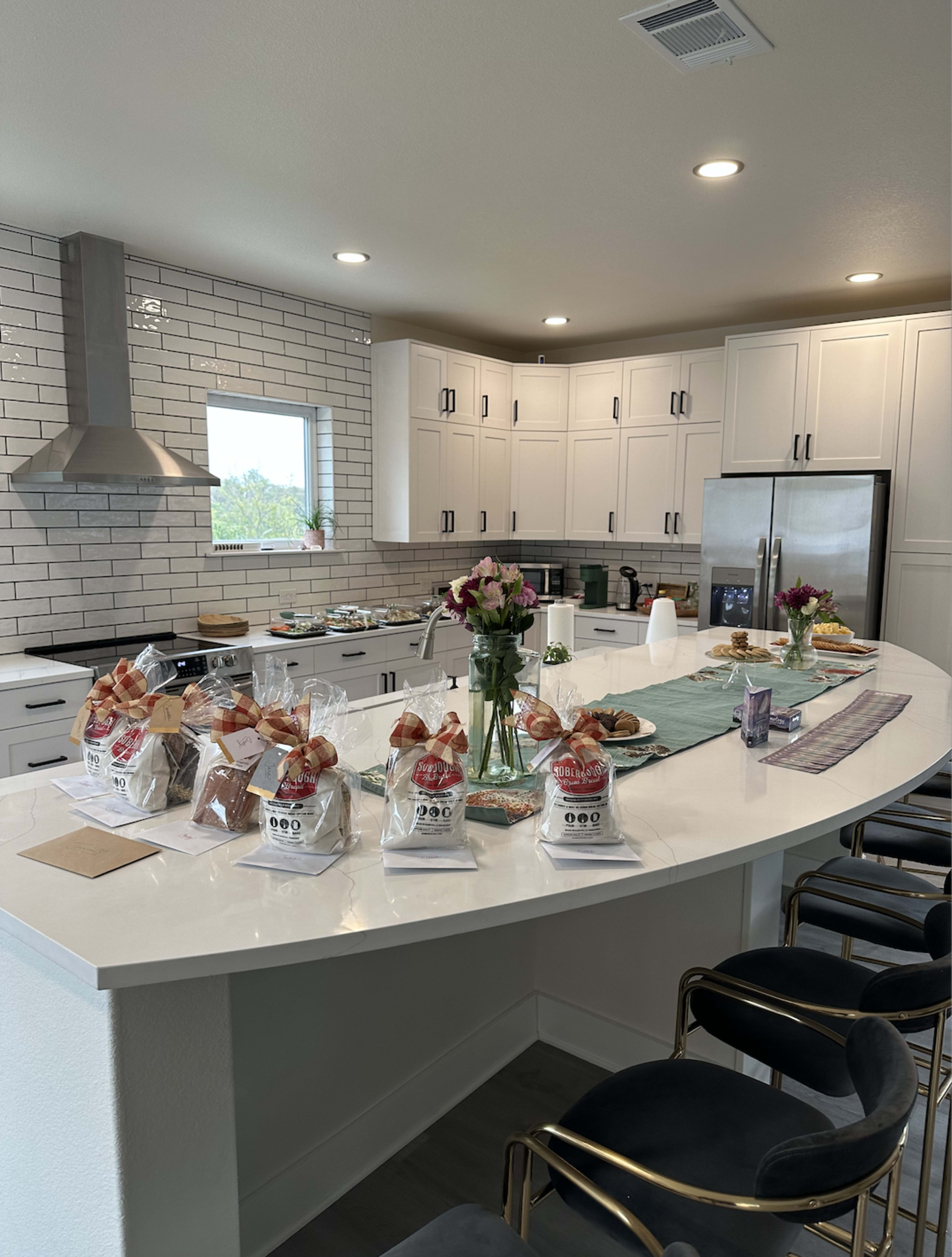 A modern kitchen with a white island countertop features several gift bags, a vase of flowers, and various snacks arranged on a green table runner.