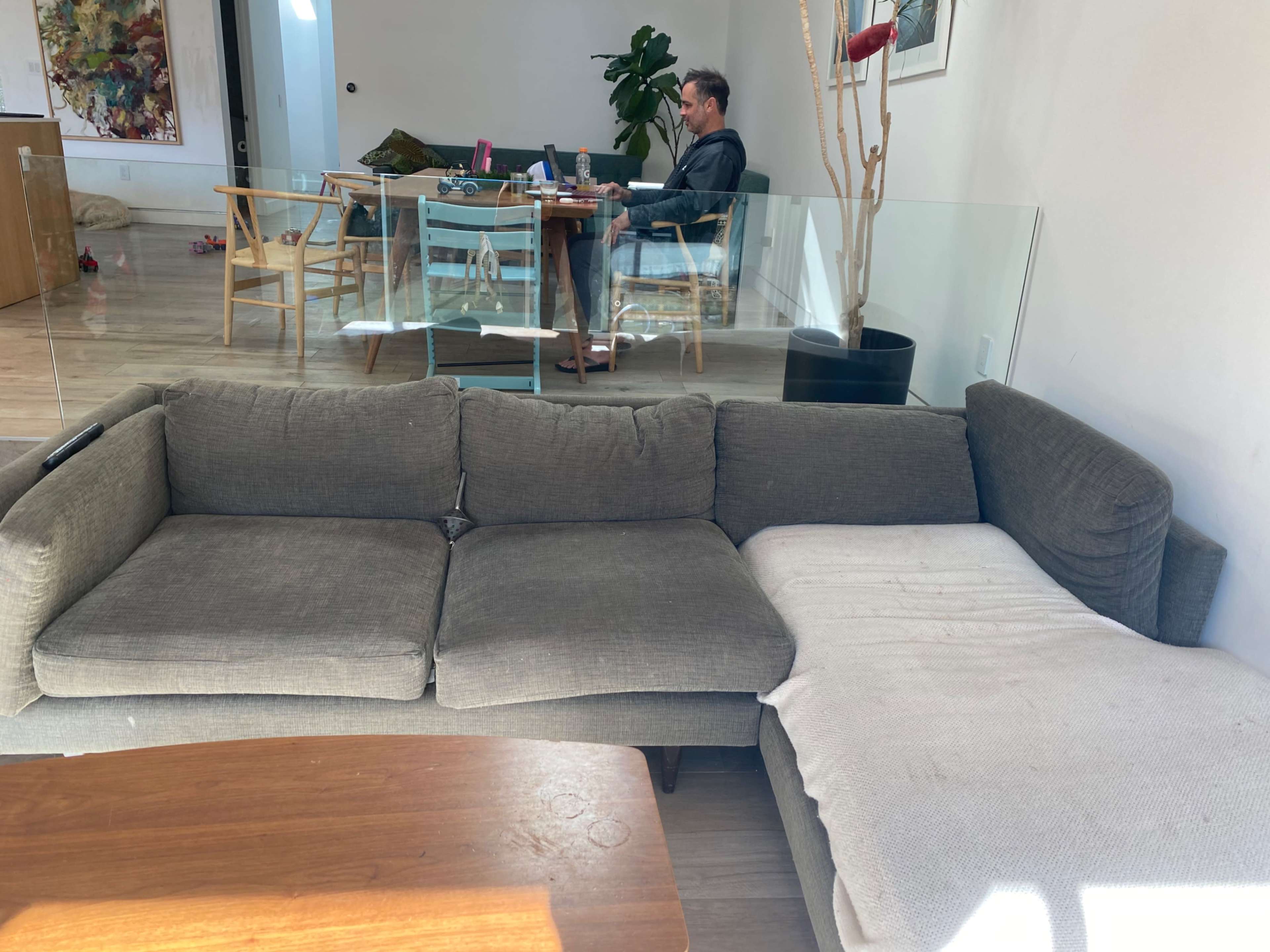 A man sits at a table in the background while a sectional sofa with a white blanket drapes over one side is in the foreground.