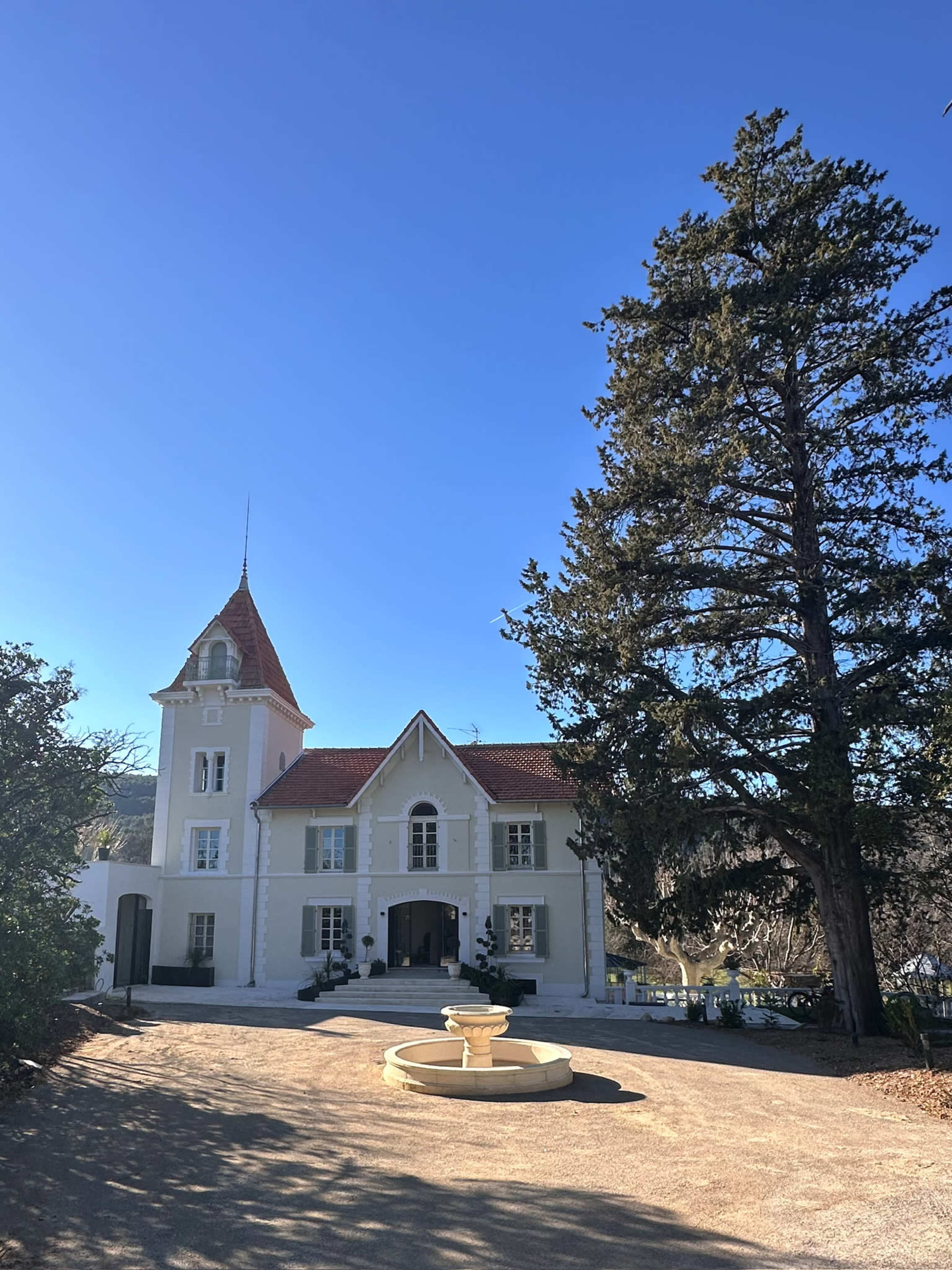 A large, white house with a pointed roof and prominent tower stands at the end of a gravel driveway, accompanied by a fountain and tall trees.