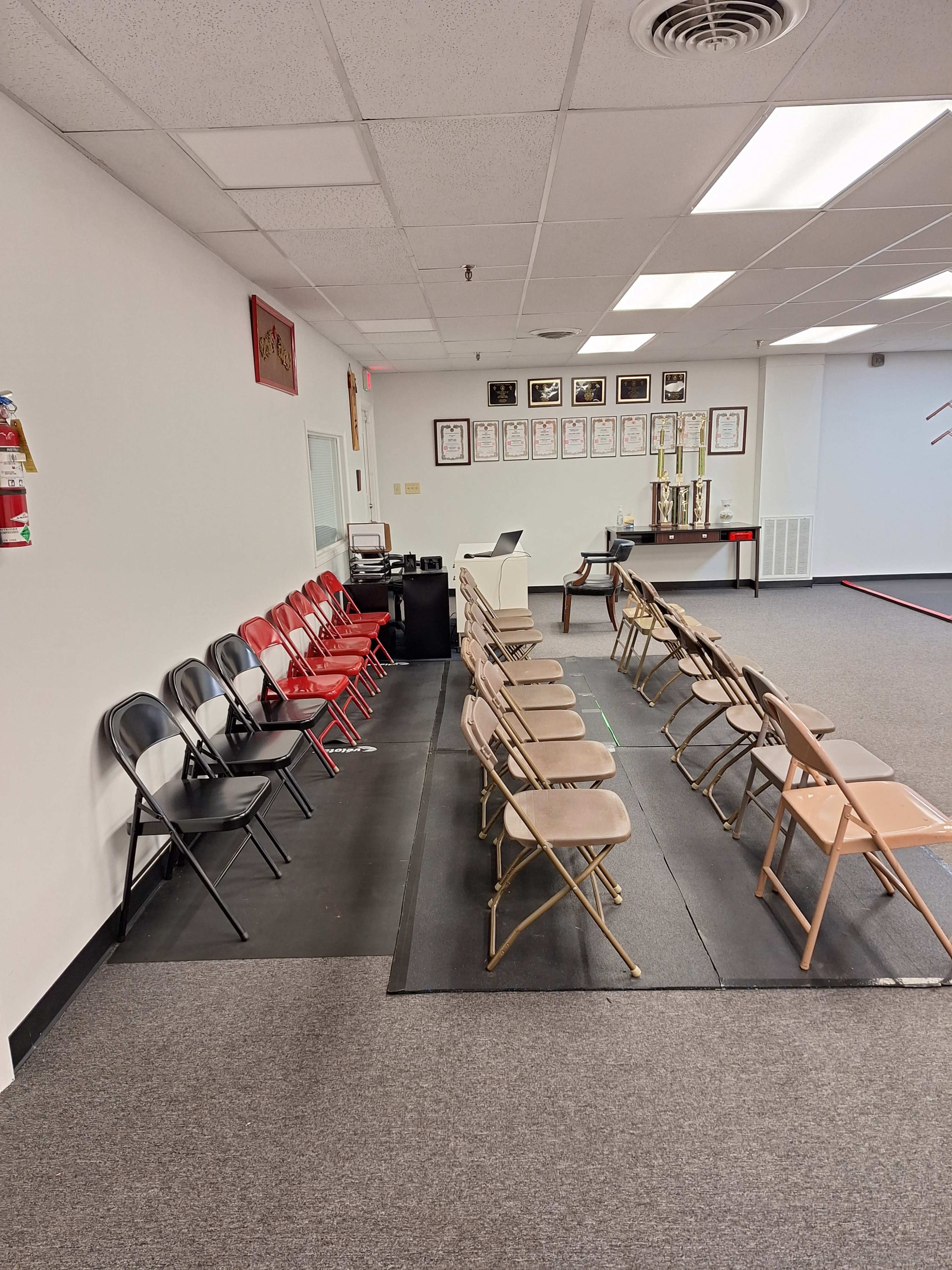 The image shows a row of red and black folding chairs arranged on a gray floor inside a spacious room with white walls and a visible trophy display.