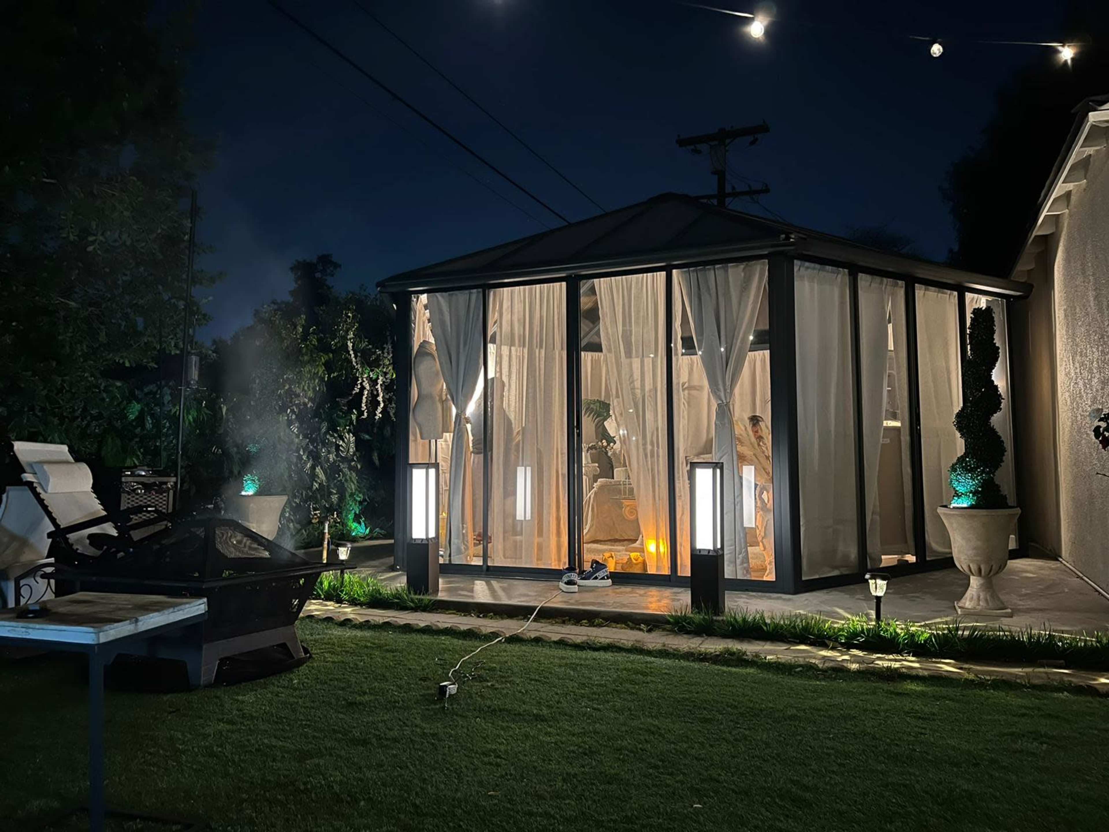 A glass-enclosed structure with sheer curtains is illuminated at night, surrounded by greenery and outdoor lights.