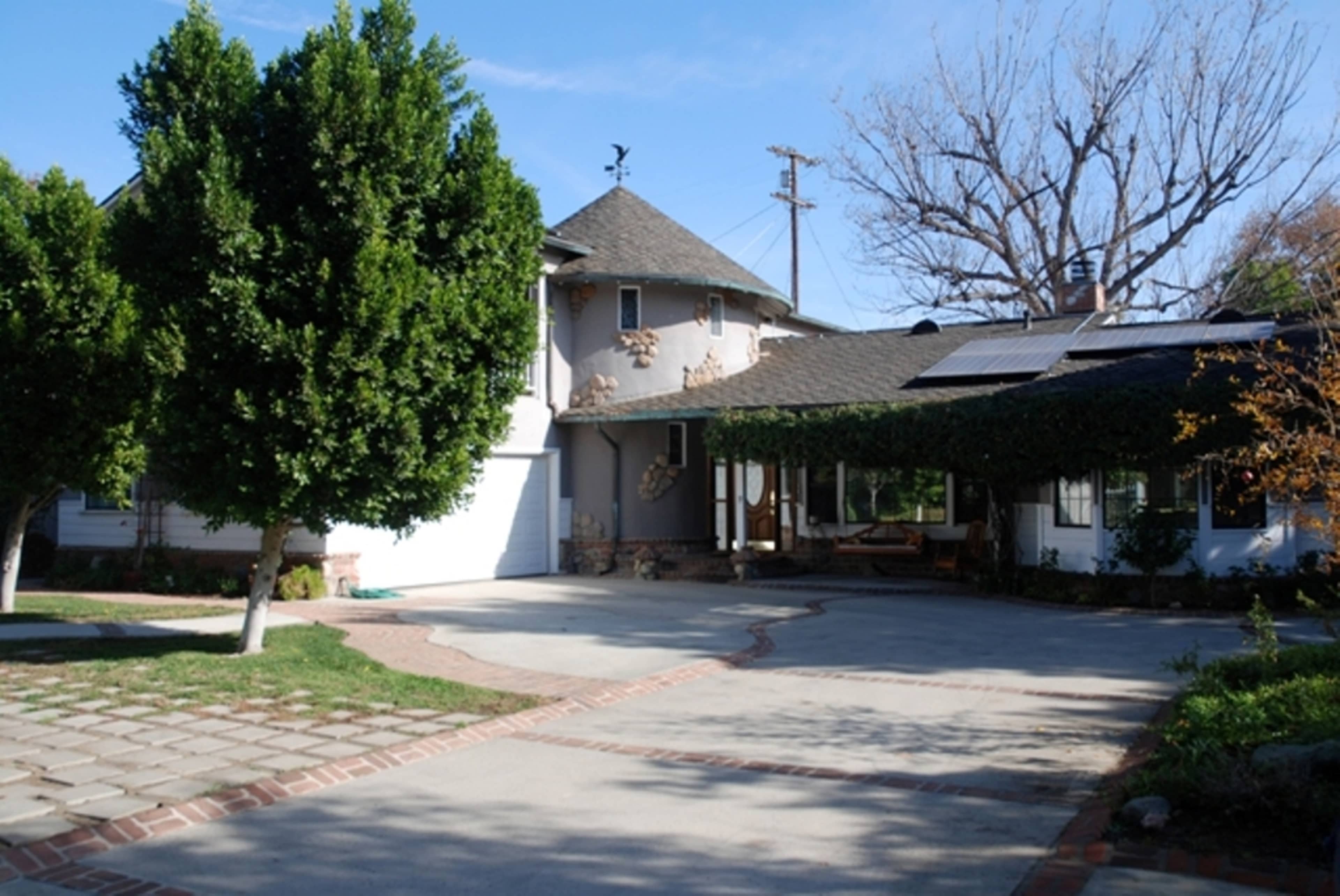 A multi-story house with a circular front entryway, a driveway made of concrete and bricks, surrounded by well-maintained trees, and a clear blue sky.