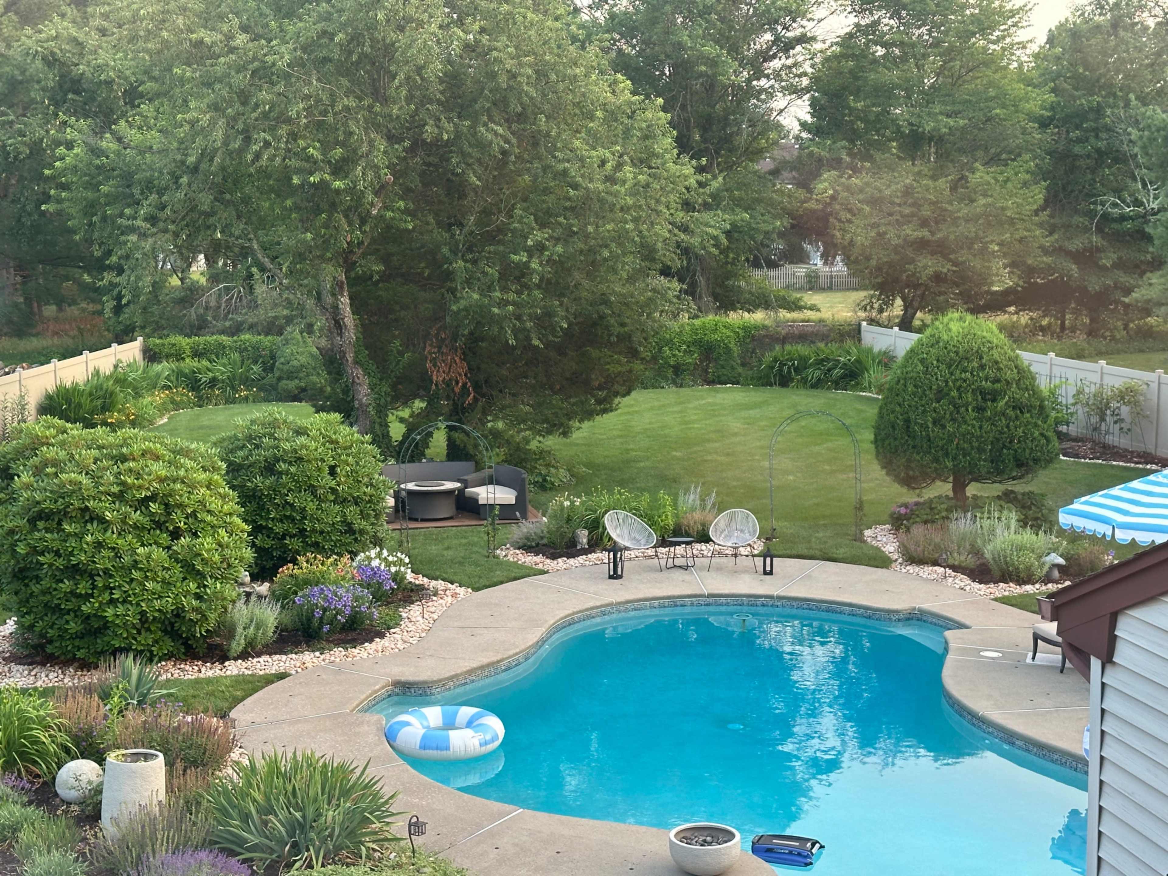 The image shows a residential backyard with a swimming pool surrounded by greenery and outdoor seating.