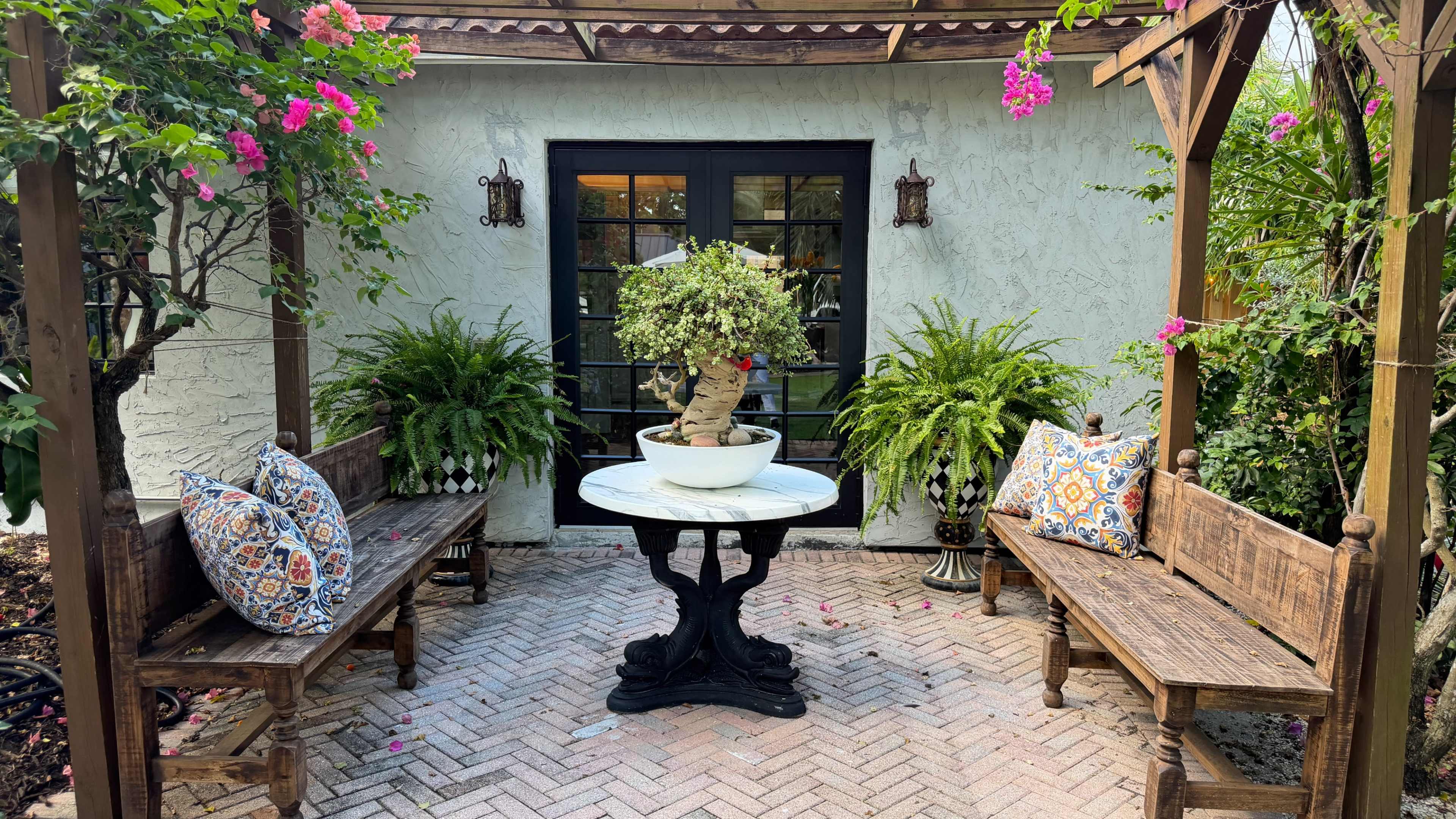The image shows a small outdoor seating area with two wooden benches, a round table, and a central potted plant surrounded by lush greenery and flowers.