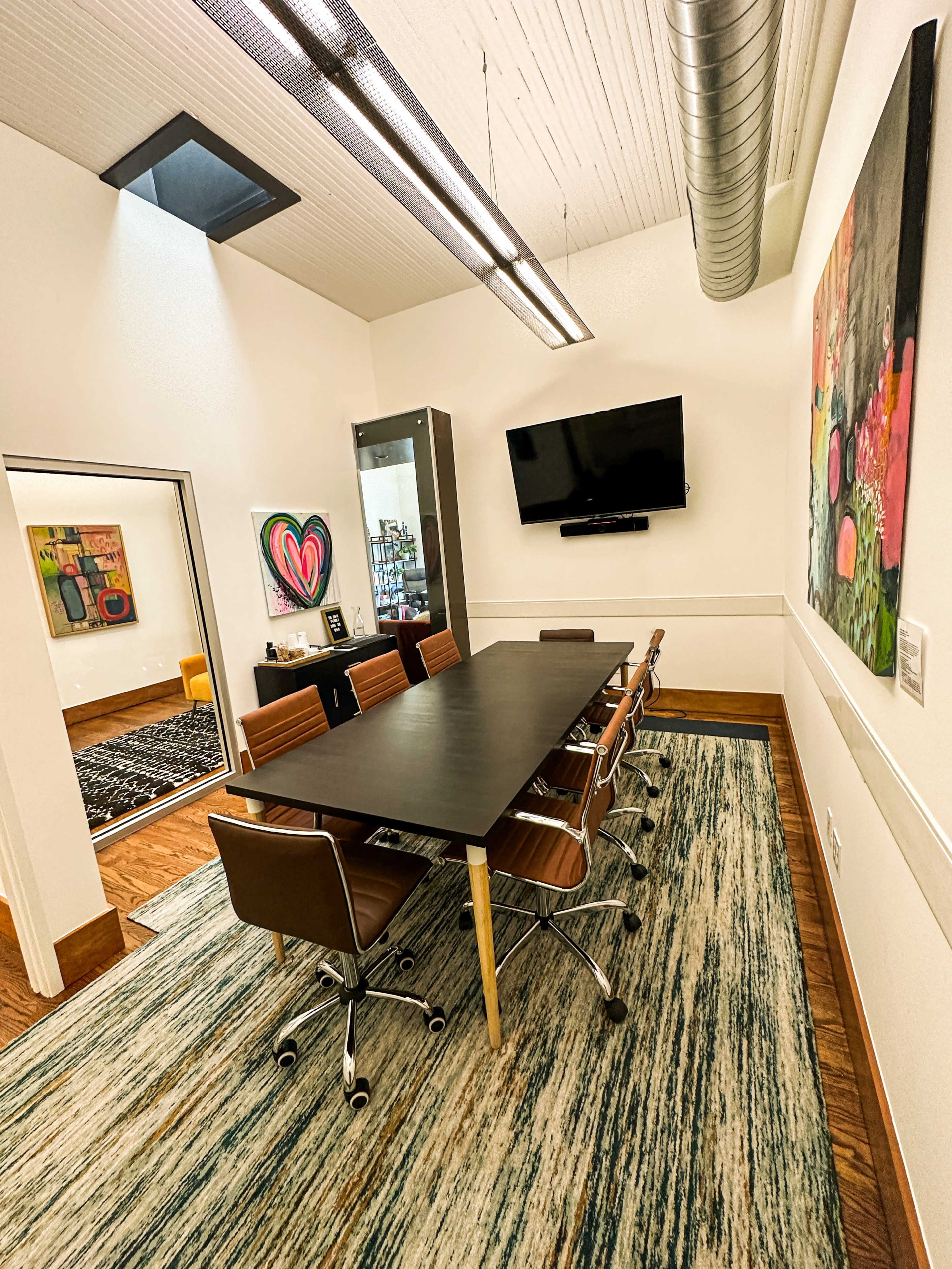 The image shows a modern conference room with a long table and six chairs, a wall-mounted TV, and colorful artworks on the walls.