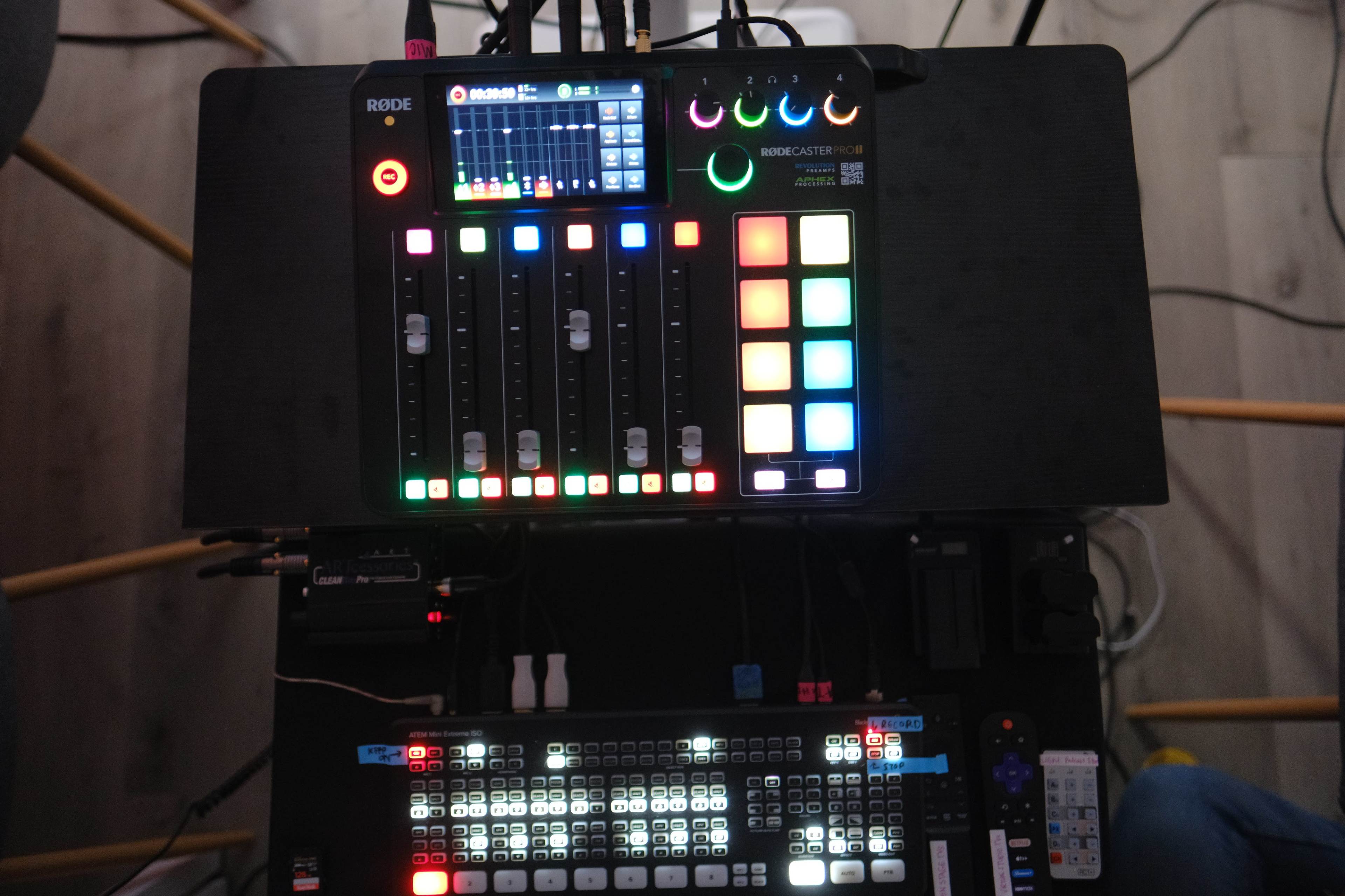 The image shows a digital audio mixer with colorful buttons and sliders, positioned on a desk alongside a control pad.