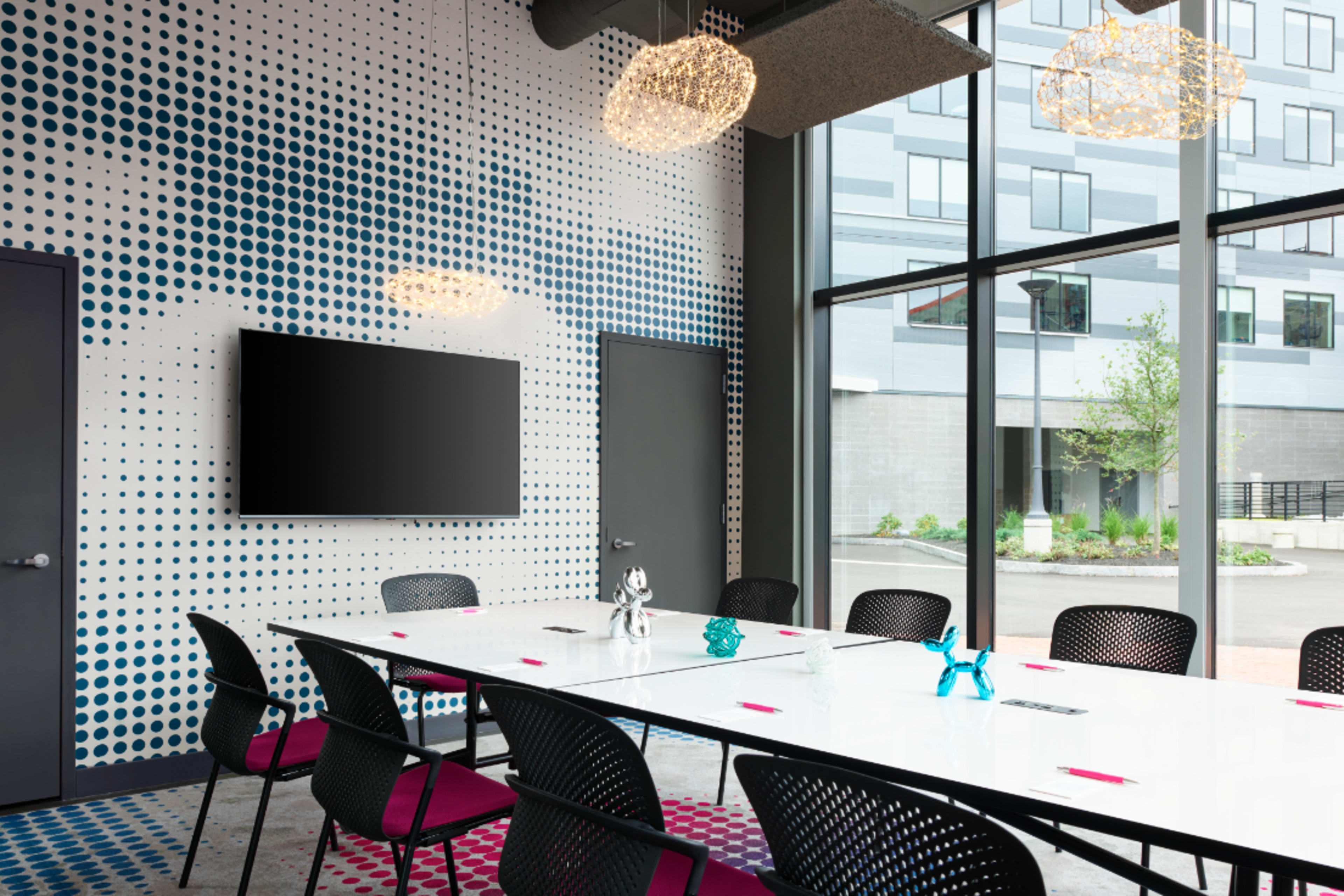 A modern conference room features a long white table surrounded by black chairs, with a large window displaying an outdoor view and a wall decorated with blue polka dots.