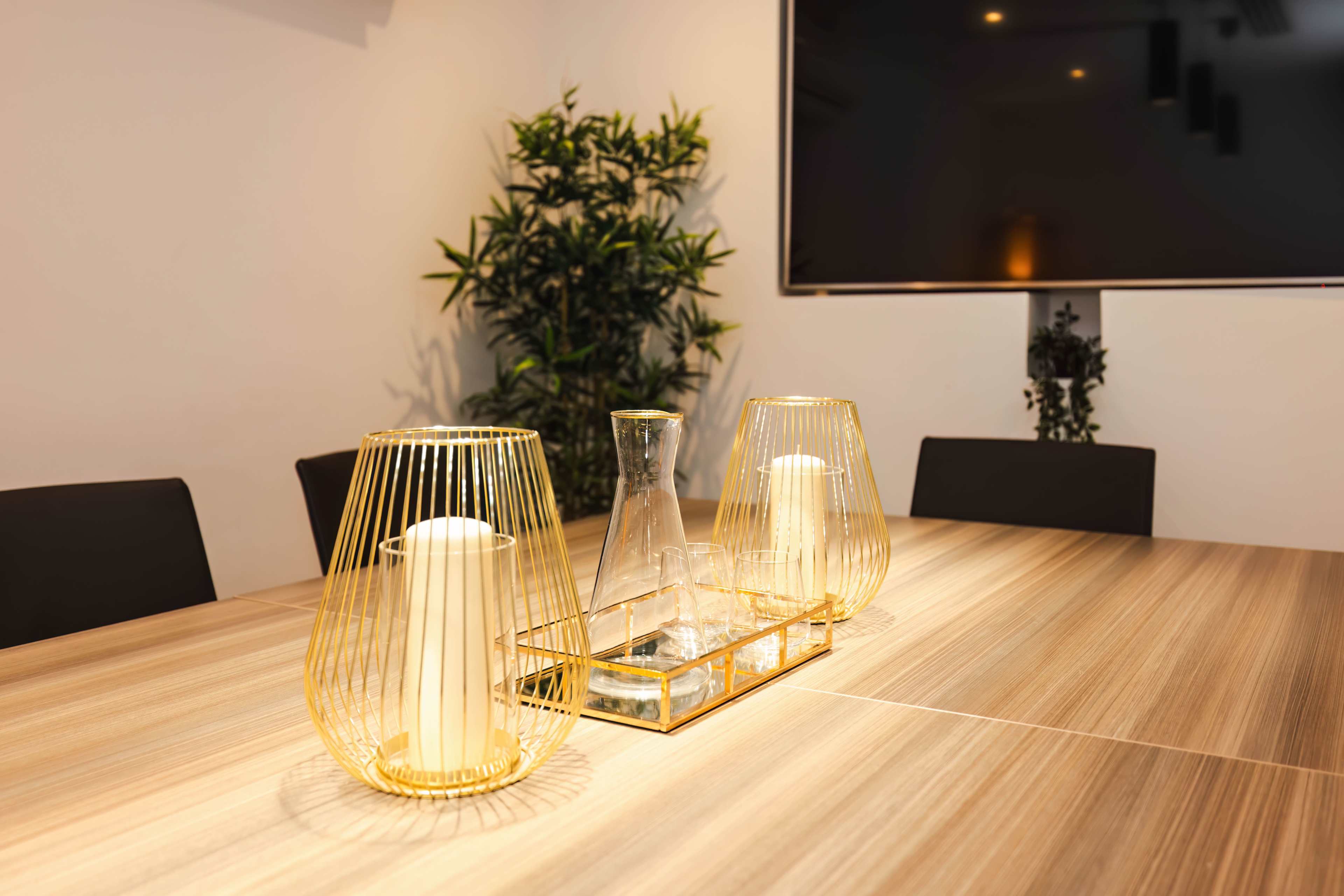 A modern dining table is set with gold wire candle holders, a glass carafe, and a decorative tray, surrounded by black chairs and greenery.