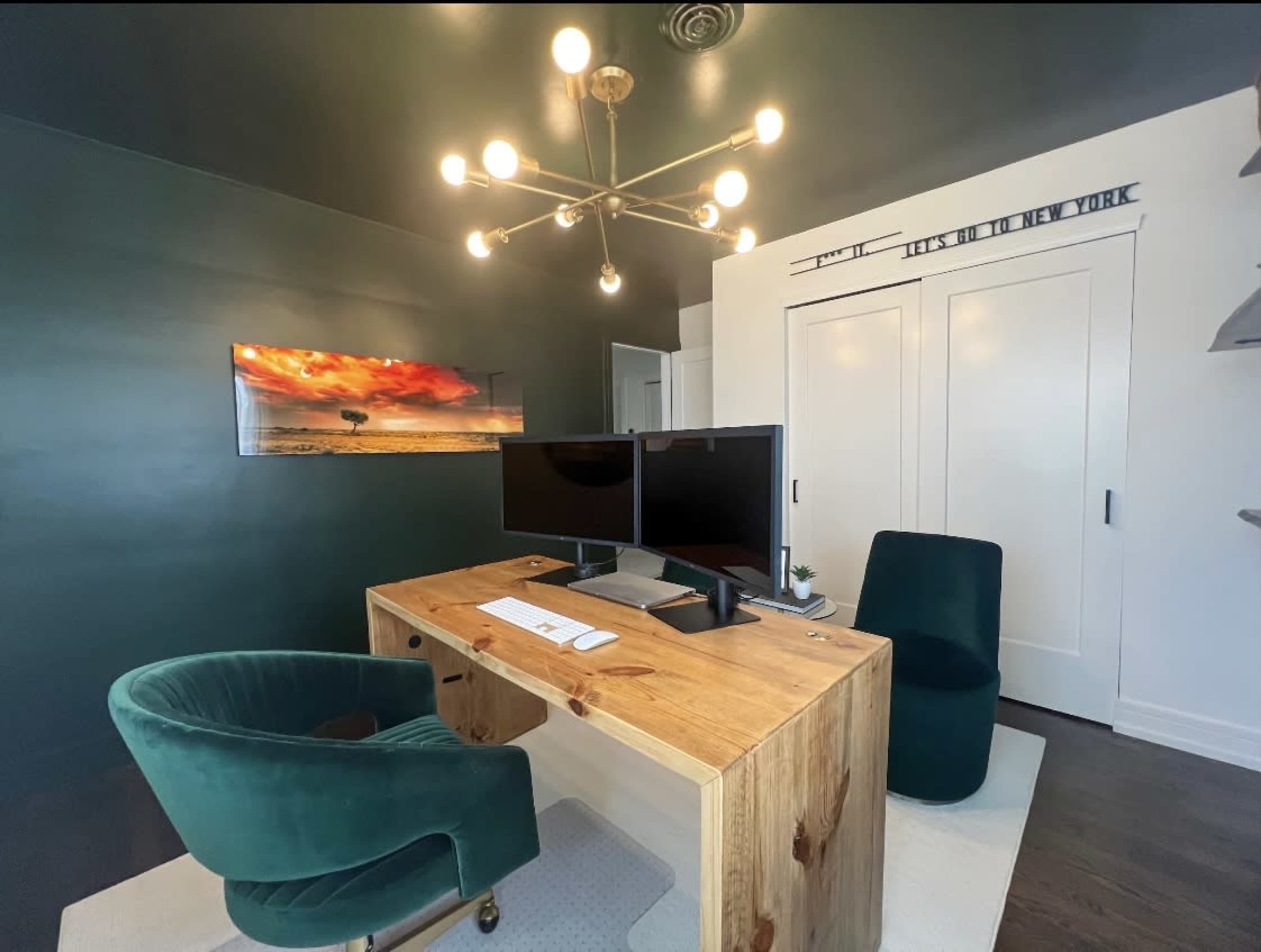 A modern office space features a wooden desk with two monitors, a green velvet chair, and a white carpet against a dark green wall, with a landscape painting and a closed closet in the background.