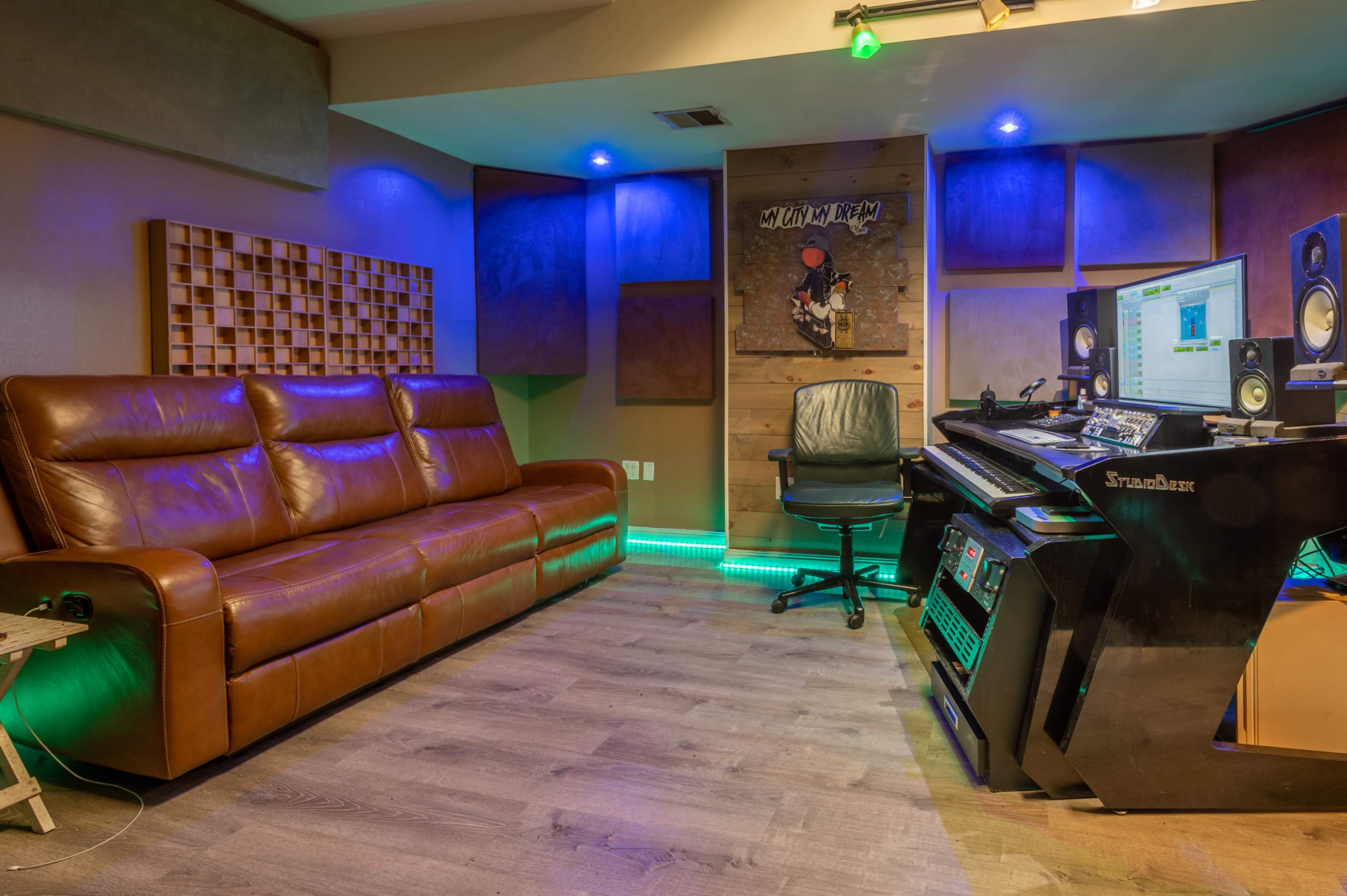 The image shows a modern music studio with a leather couch, computer desk, and acoustic paneling, illuminated by colored lights.