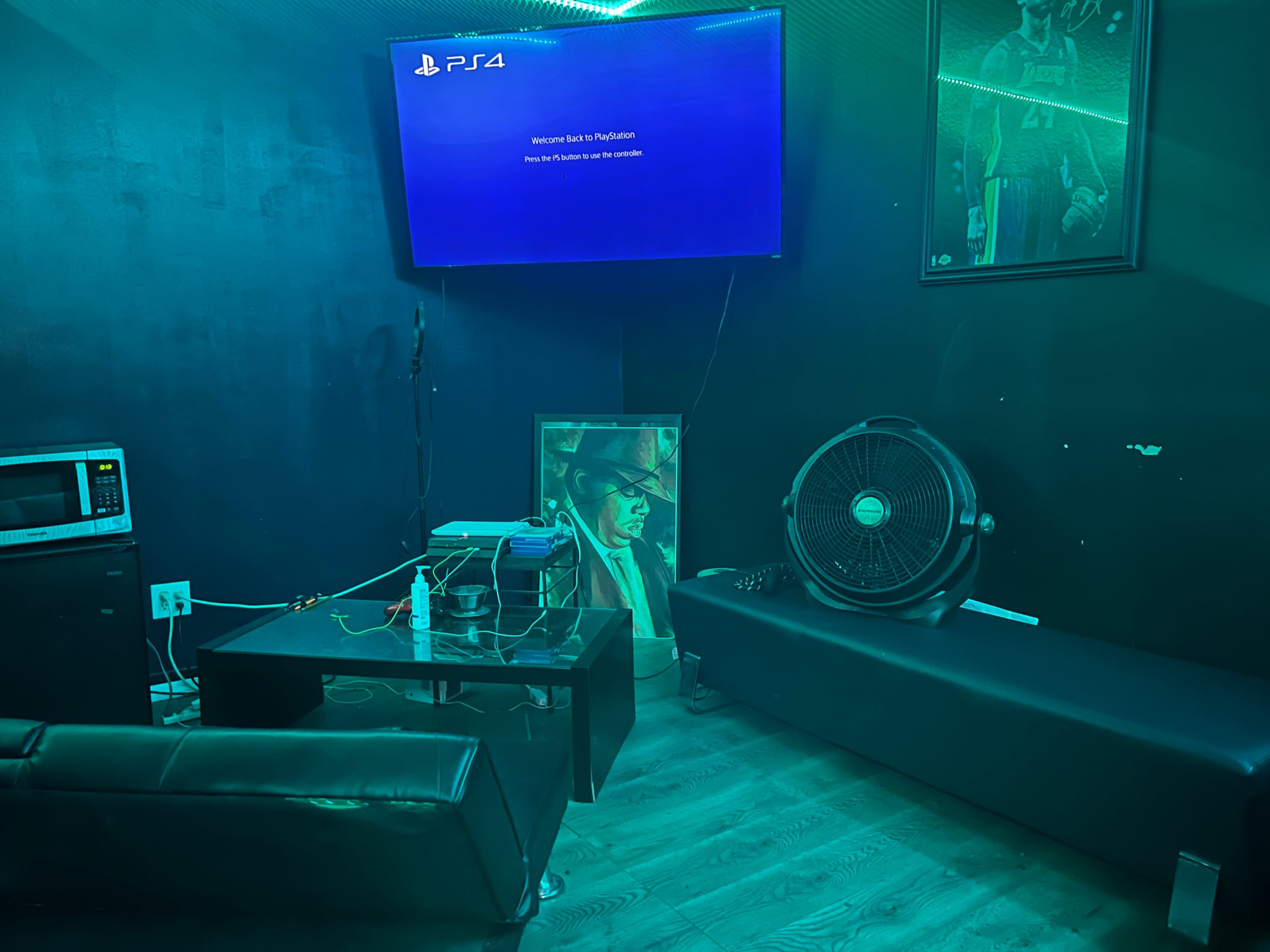 The image shows a dimly lit room featuring a PlayStation 4 console connected to a TV, a black sofa, a fan, a microwave, and a framed portrait on the floor.