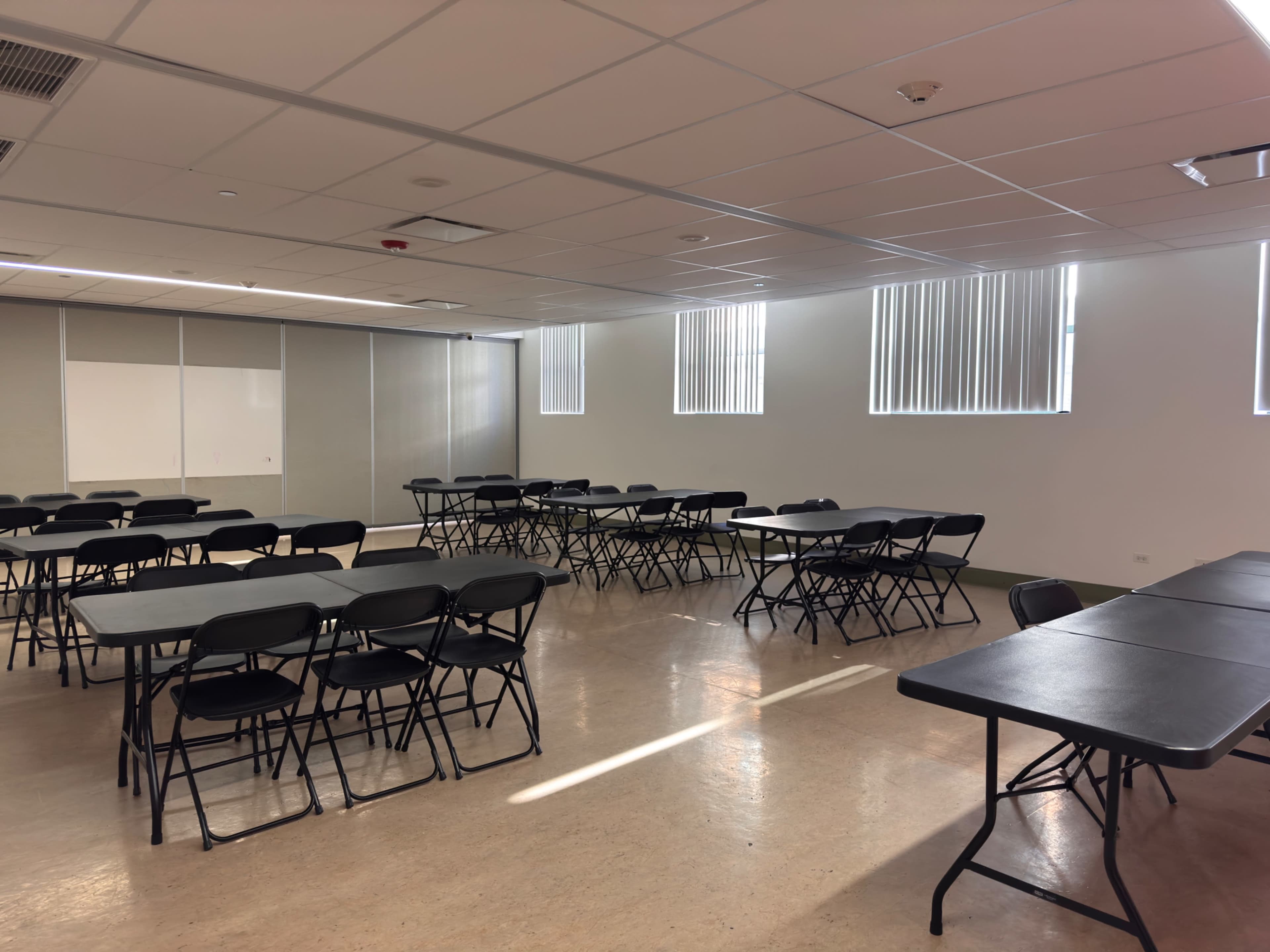 The image shows a classroom or meeting space with multiple black folding tables arranged in rows and large windows allowing natural light to enter.