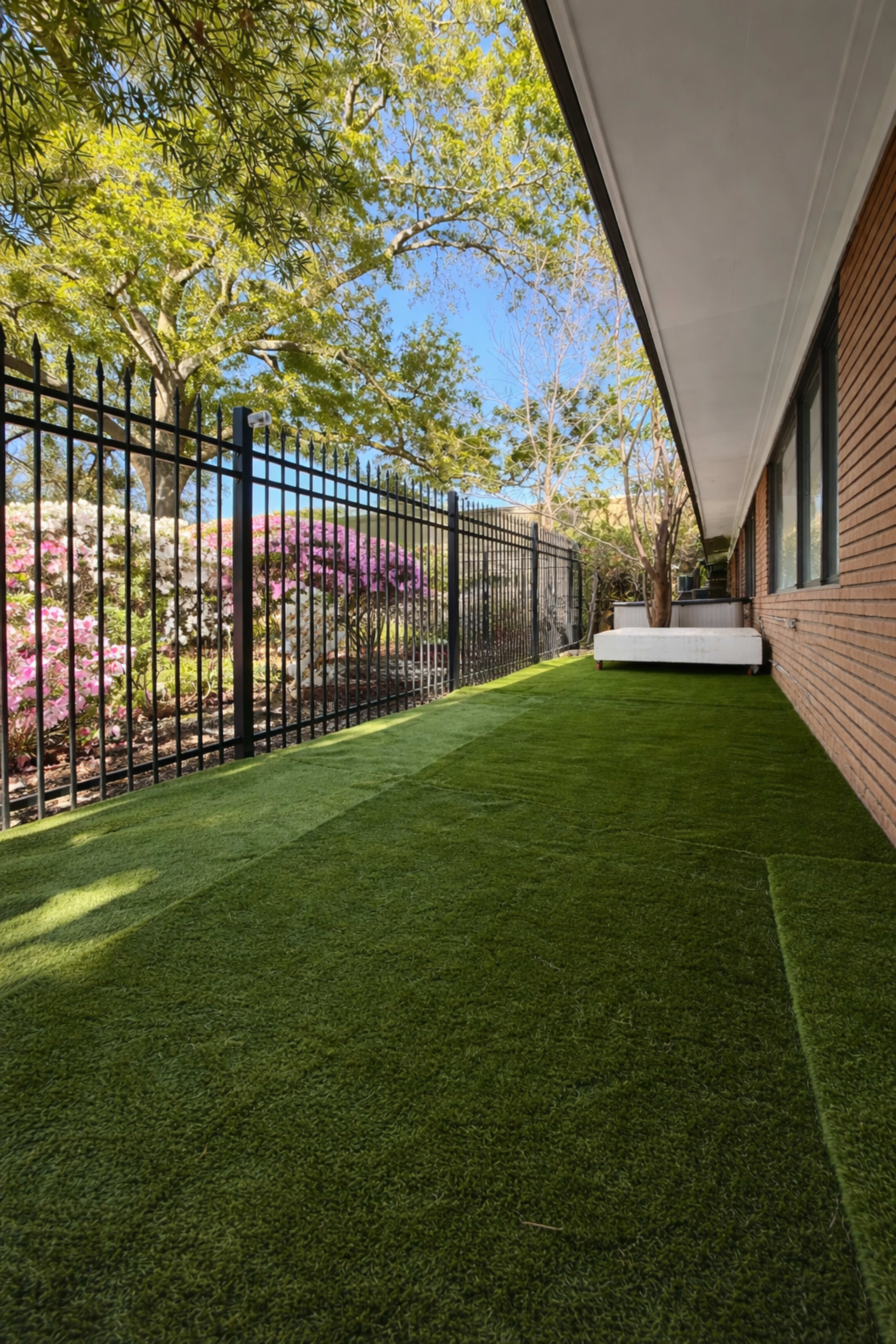 The image shows a narrow outdoor area with artificial grass, bordered by a black metal fence and colorful flowering bushes in the background.