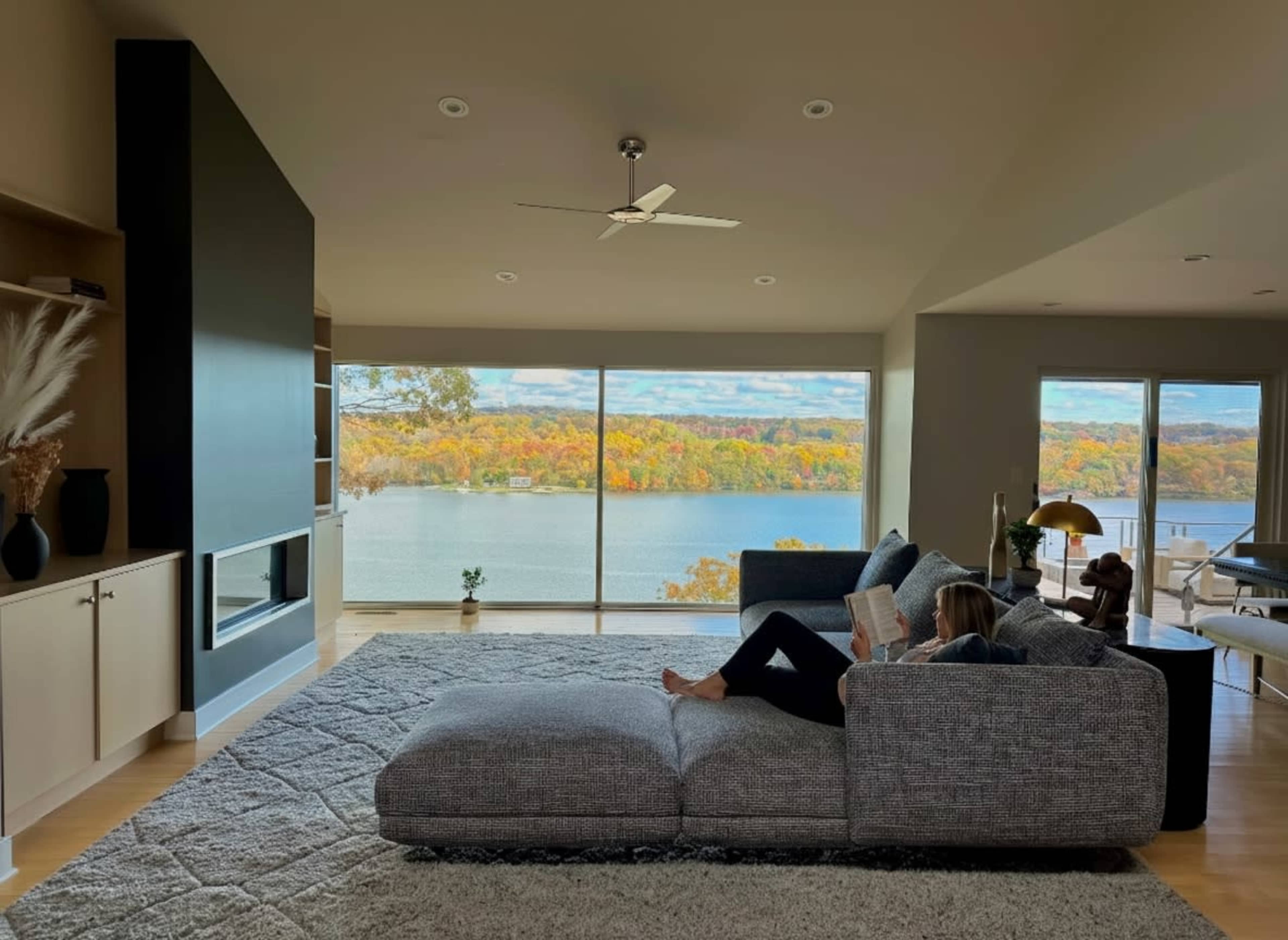 A person is seated on a large sectional sofa, reading a book while overlooking a lake surrounded by autumn-colored trees through a large window.