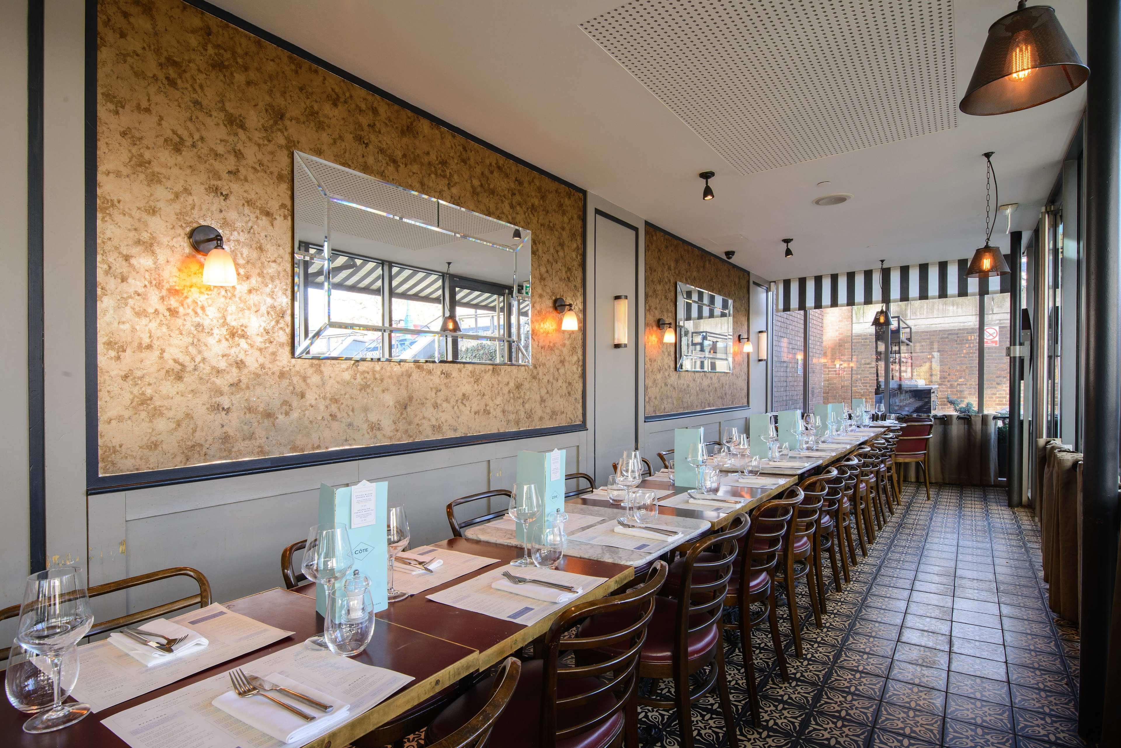The image shows a dining area of a restaurant with long tables set for guests, featuring floral wallpaper, a large mirror, and pendant lighting.
