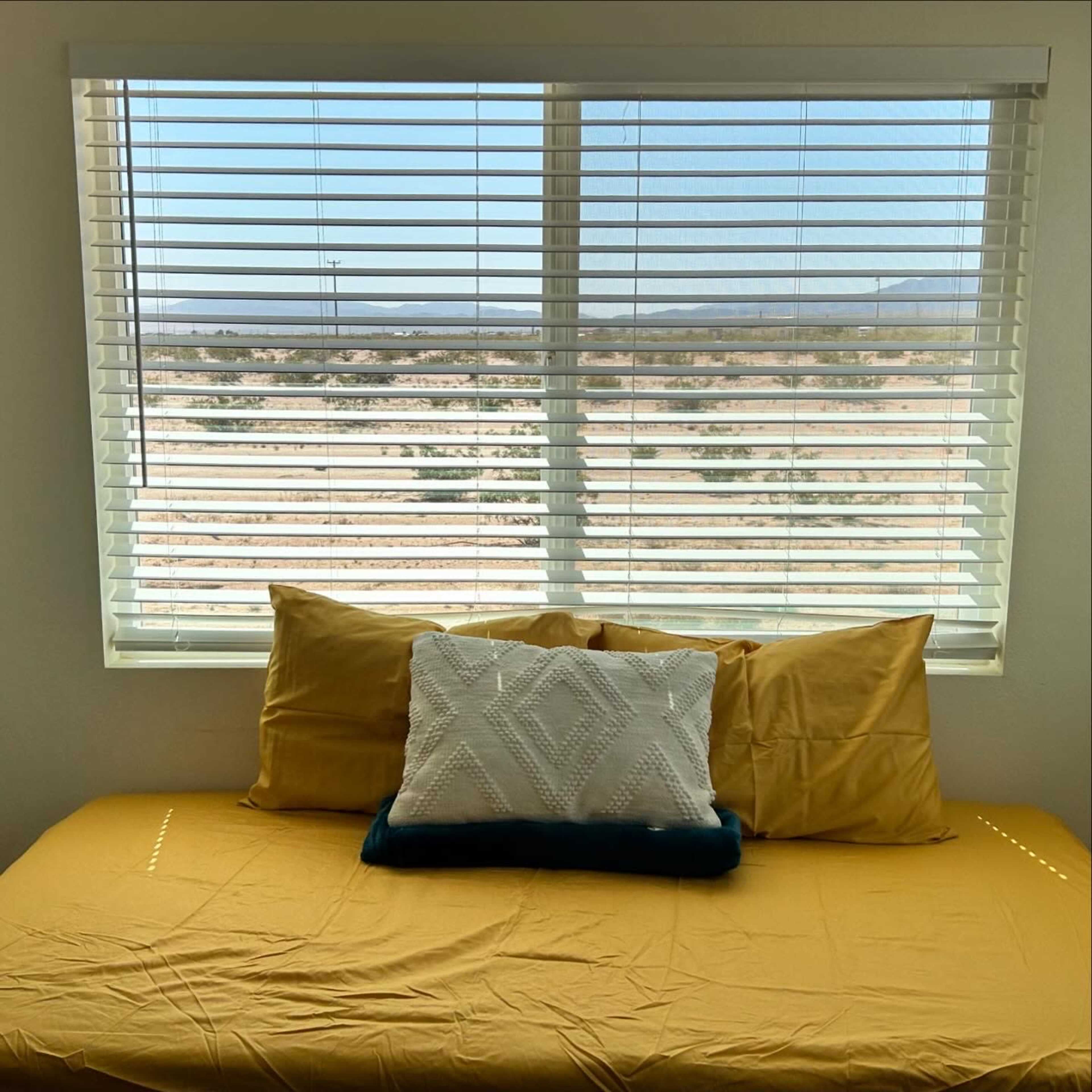 A bed with yellow bedding and a decorative pillow is positioned in front of a window with white blinds, offering a view of a desert landscape.