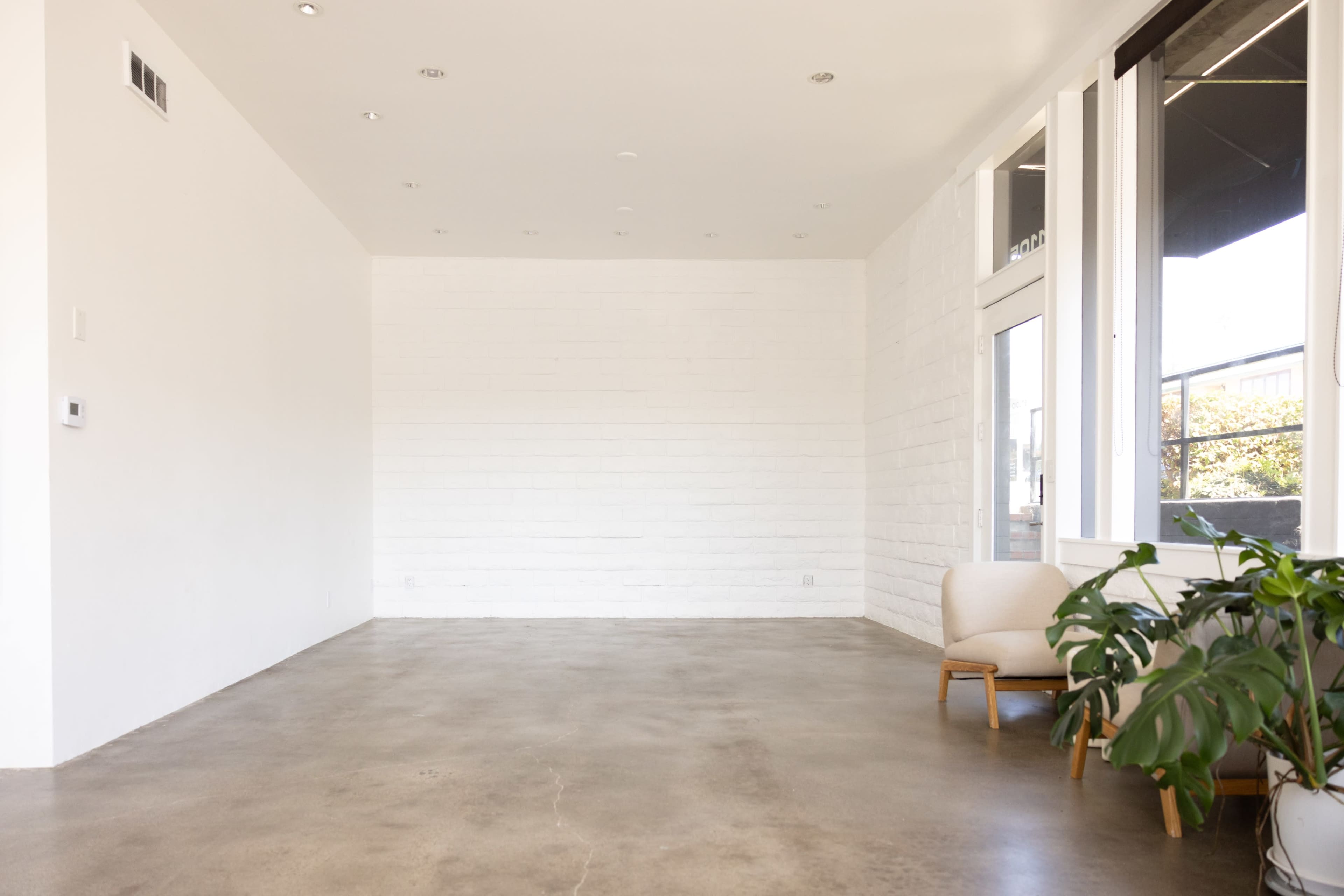 The image shows a minimalist room with a concrete floor, white walls, two windows, and a single chair next to a potted plant.