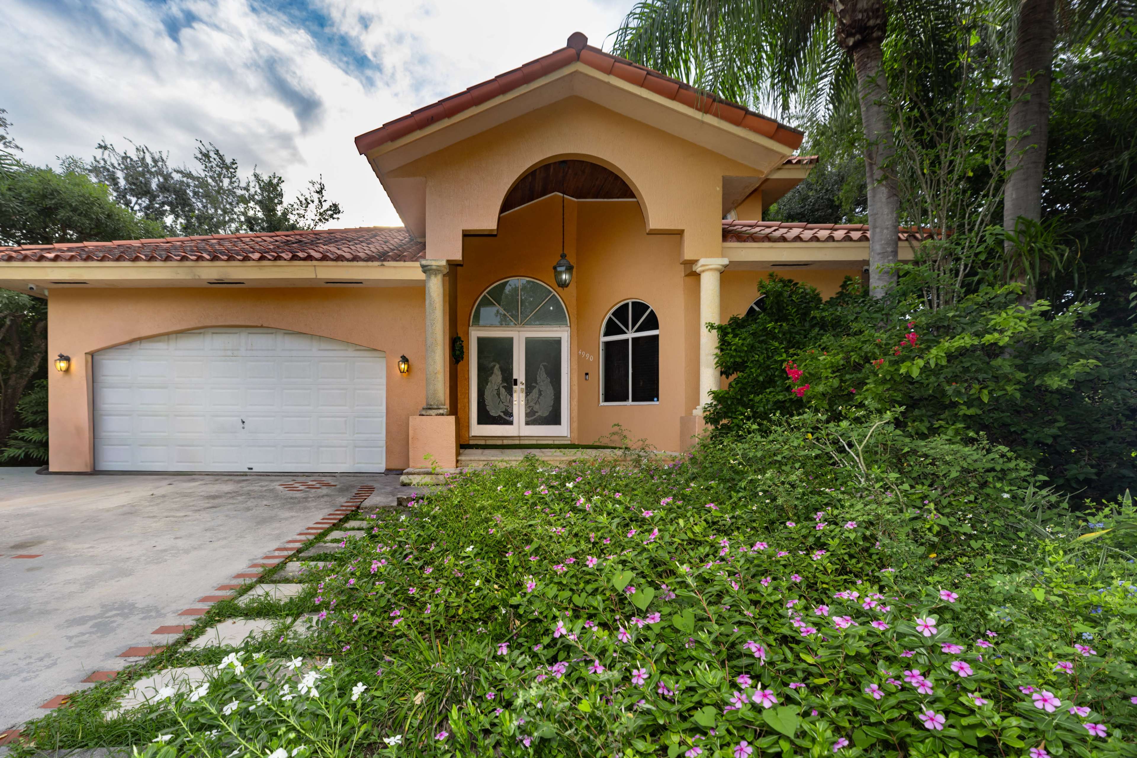 The image shows a single-story home with a light orange facade, a tiled roof, and a front yard garden featuring green foliage and pink flowers.