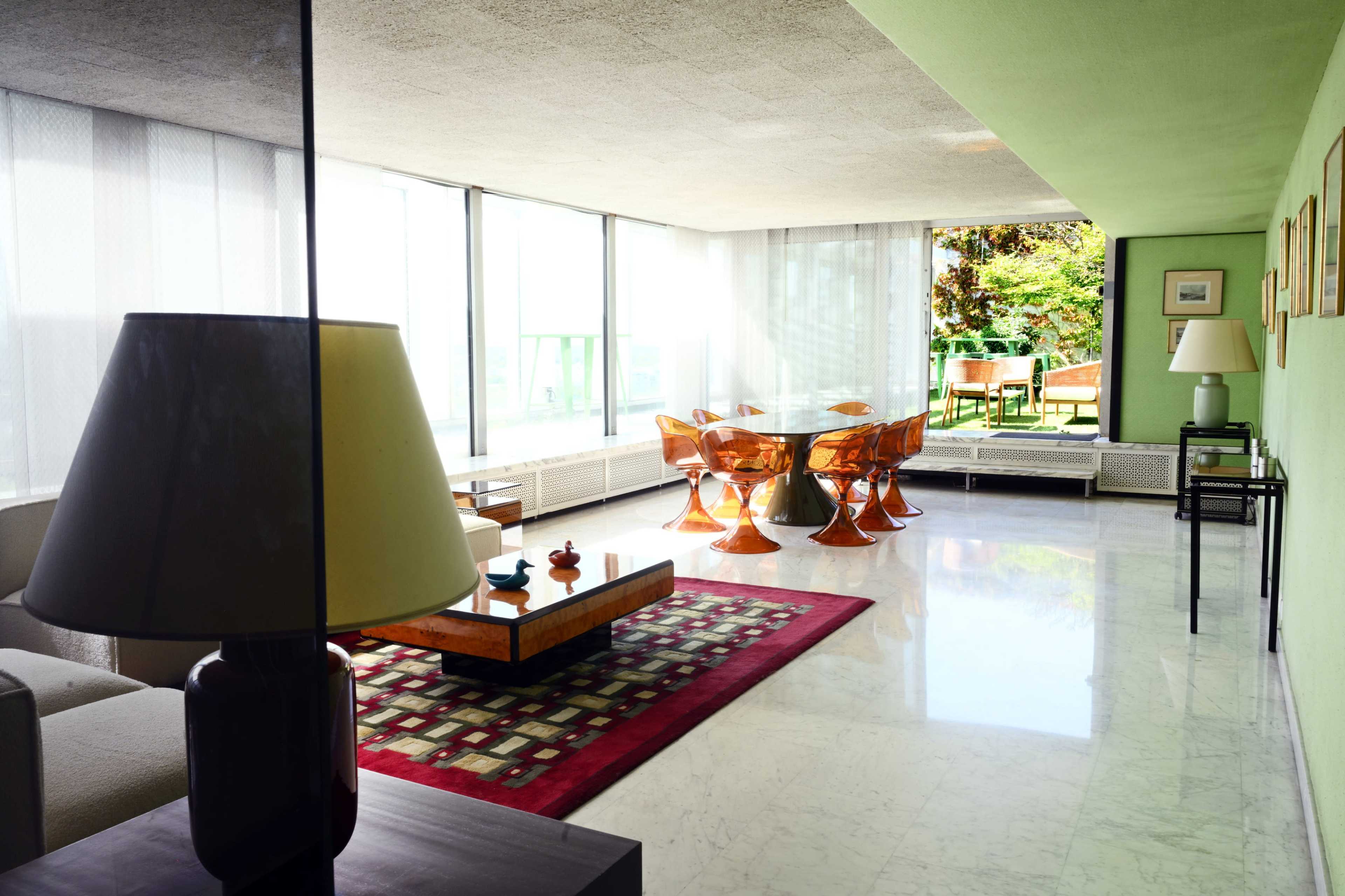 The image shows a modern living room with large windows, minimalist furniture, and a dining area featuring transparent chairs and a green wall.