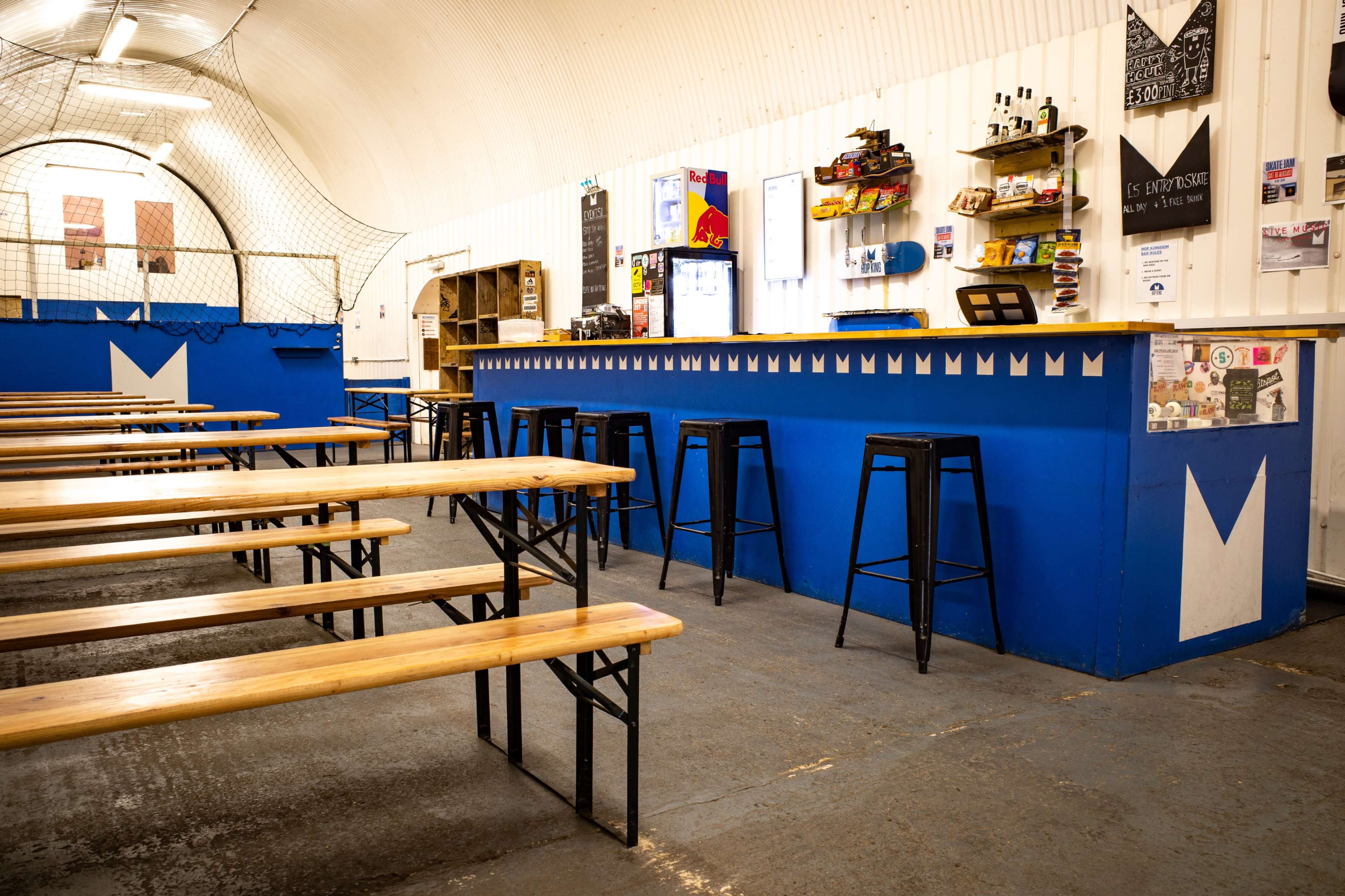 A spacious indoor bar area with wooden benches and a blue counter, featuring shelves of beverages and snacks in a well-lit environment.