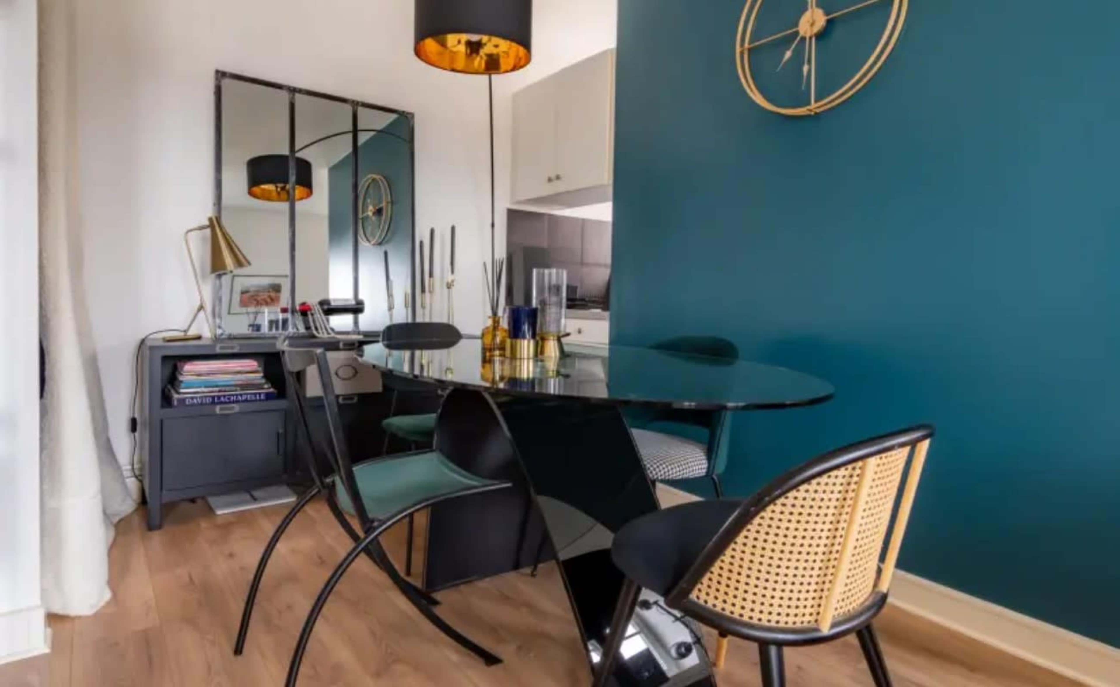 The image shows a modern dining area with a glass table, black chairs with cane backs, and a dark teal wall featuring a large clock and a mirror.