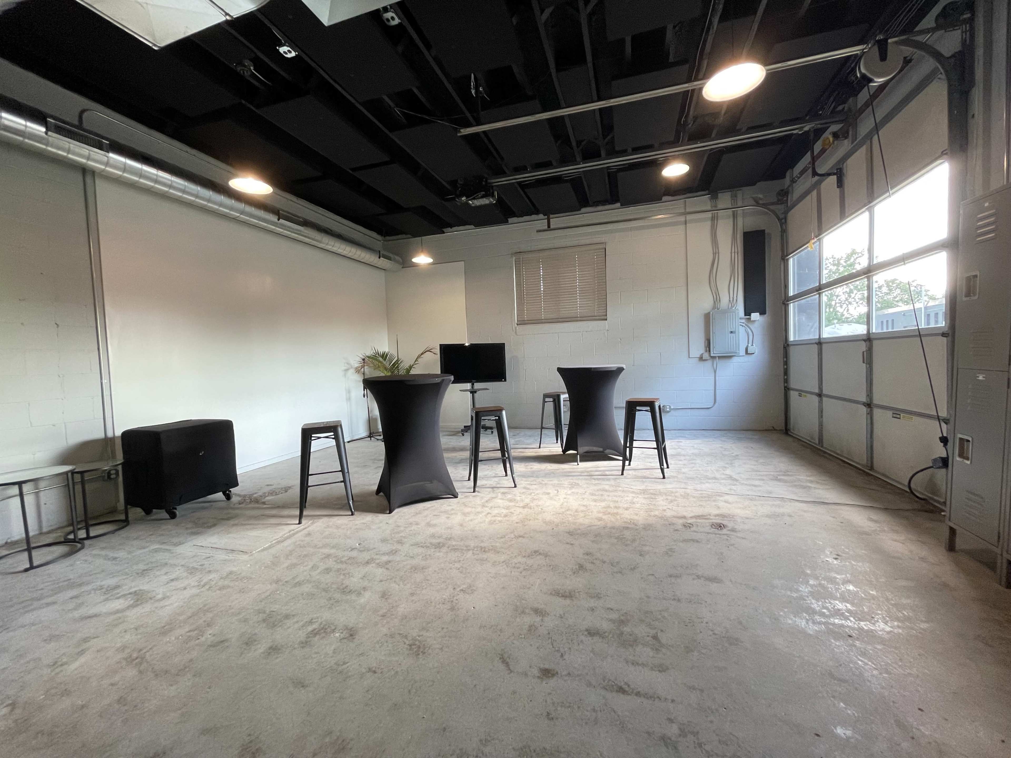An empty garage with concrete flooring, featuring three high tables and a monitor on a stand, with a large window letting in natural light.