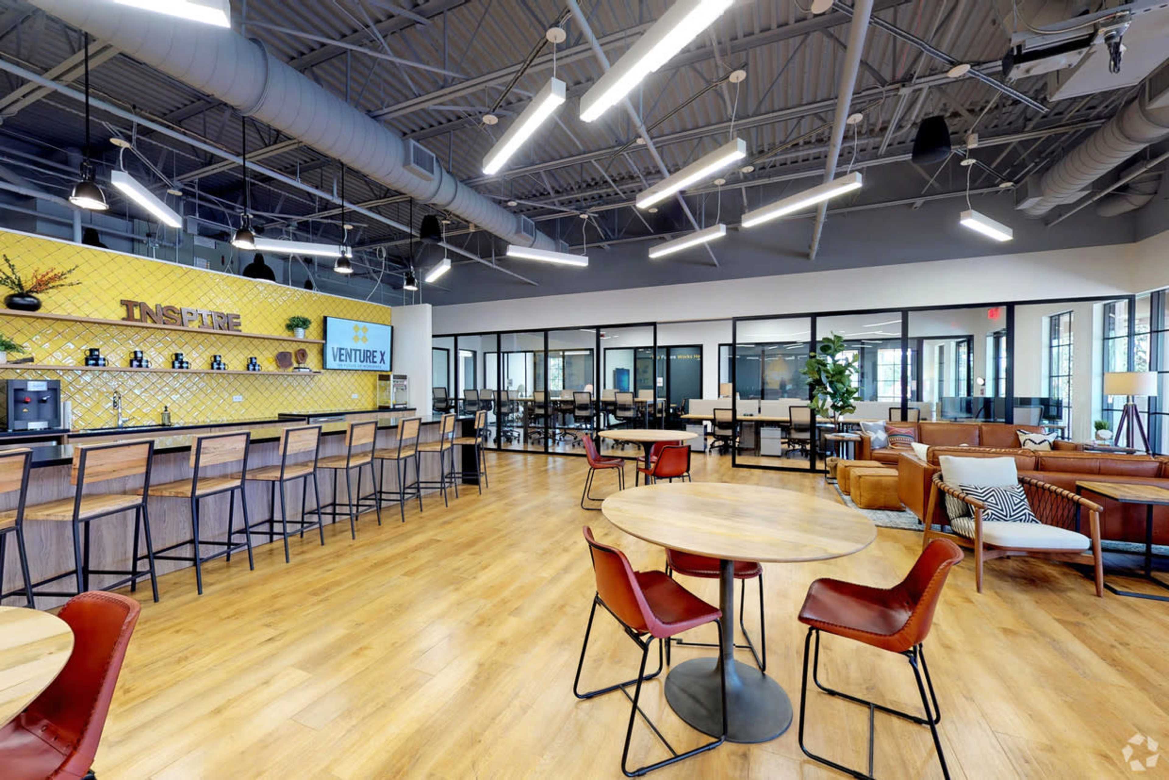 The image shows an open office space with modern furnishings, including a bar area, tables with chairs, and a lounge area, all featuring a bright and airy atmosphere.