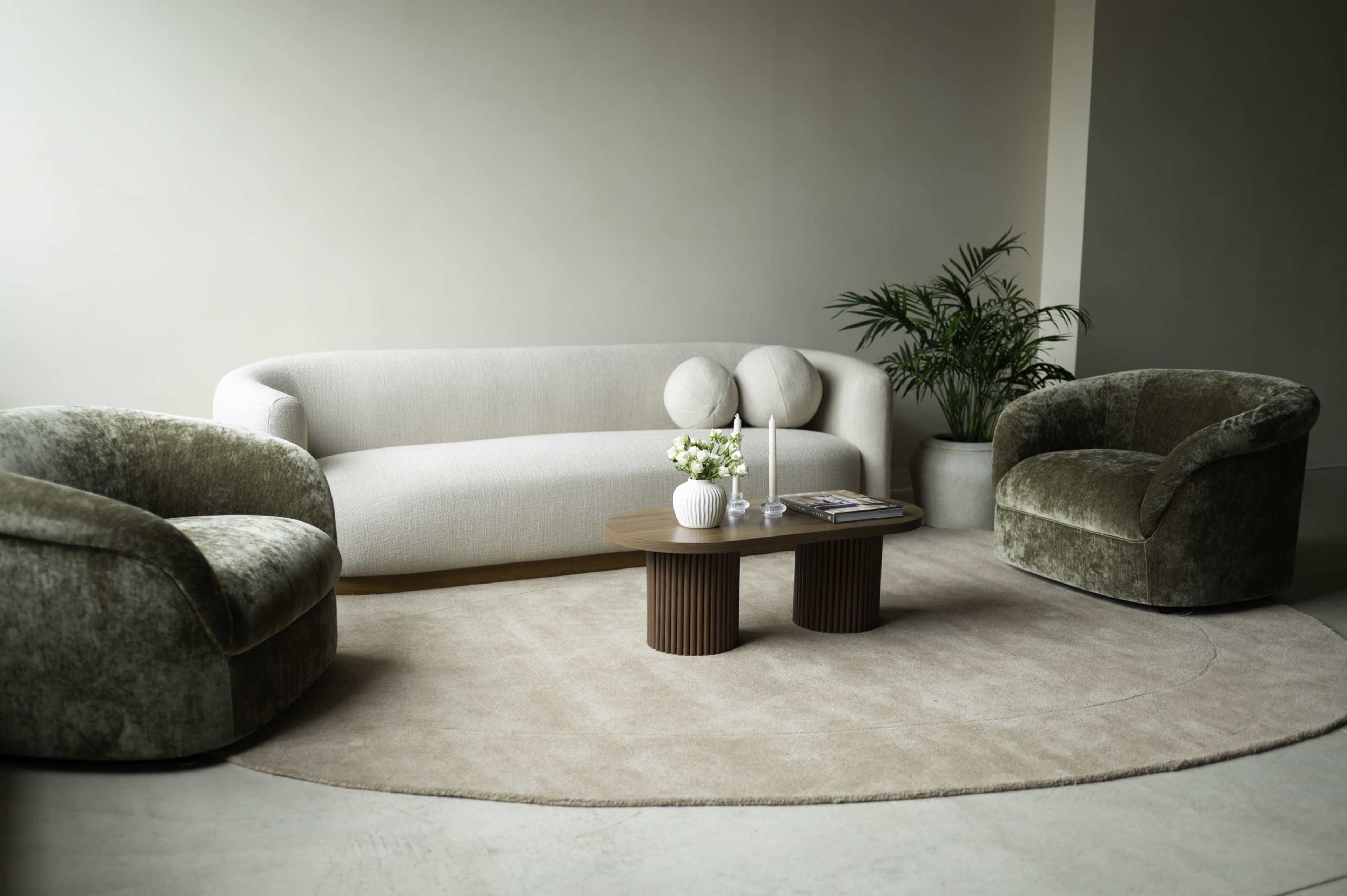 The image features a modern living room with a curved light-colored sofa, two plush green armchairs, a round rug, a small wooden coffee table, and a potted plant in the background.