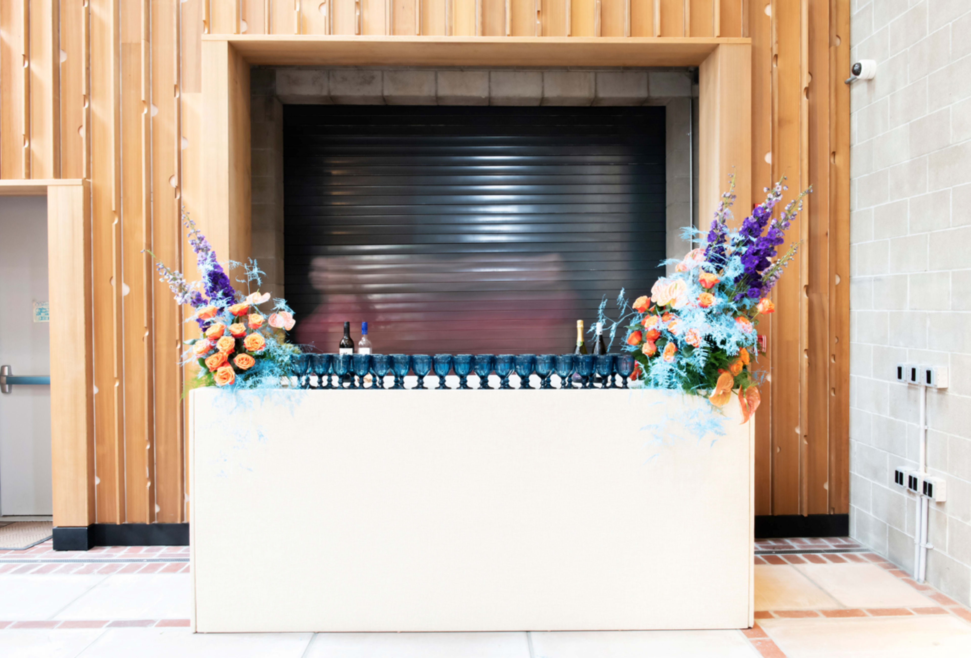 A bar counter with a variety of bottles and glasses is adorned with vibrant floral arrangements on either side, set against a wooden wall and a closed shutter.