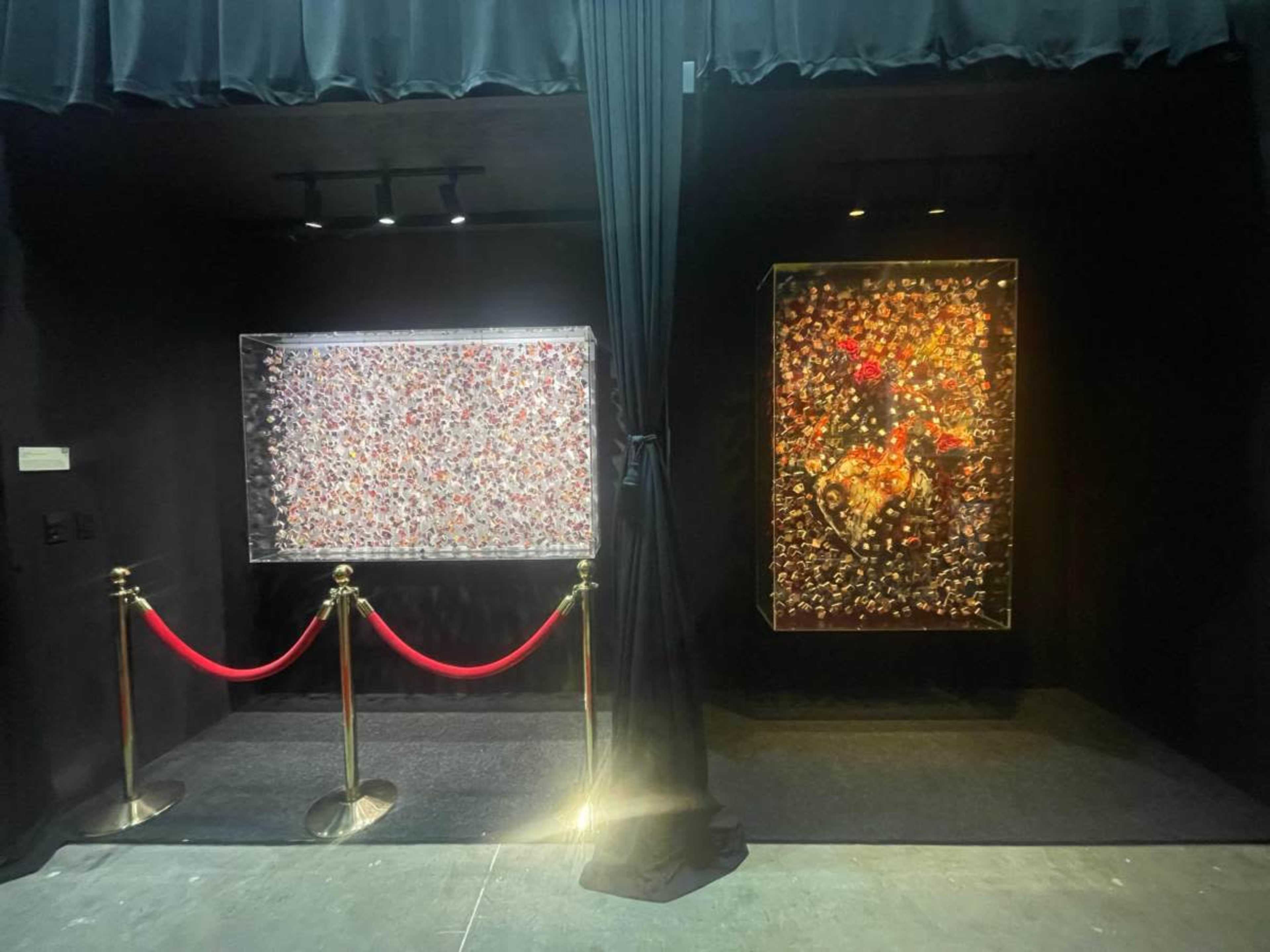 The image shows an exhibition space featuring two large, framed artworks behind a black curtain, with a red rope barrier in front.