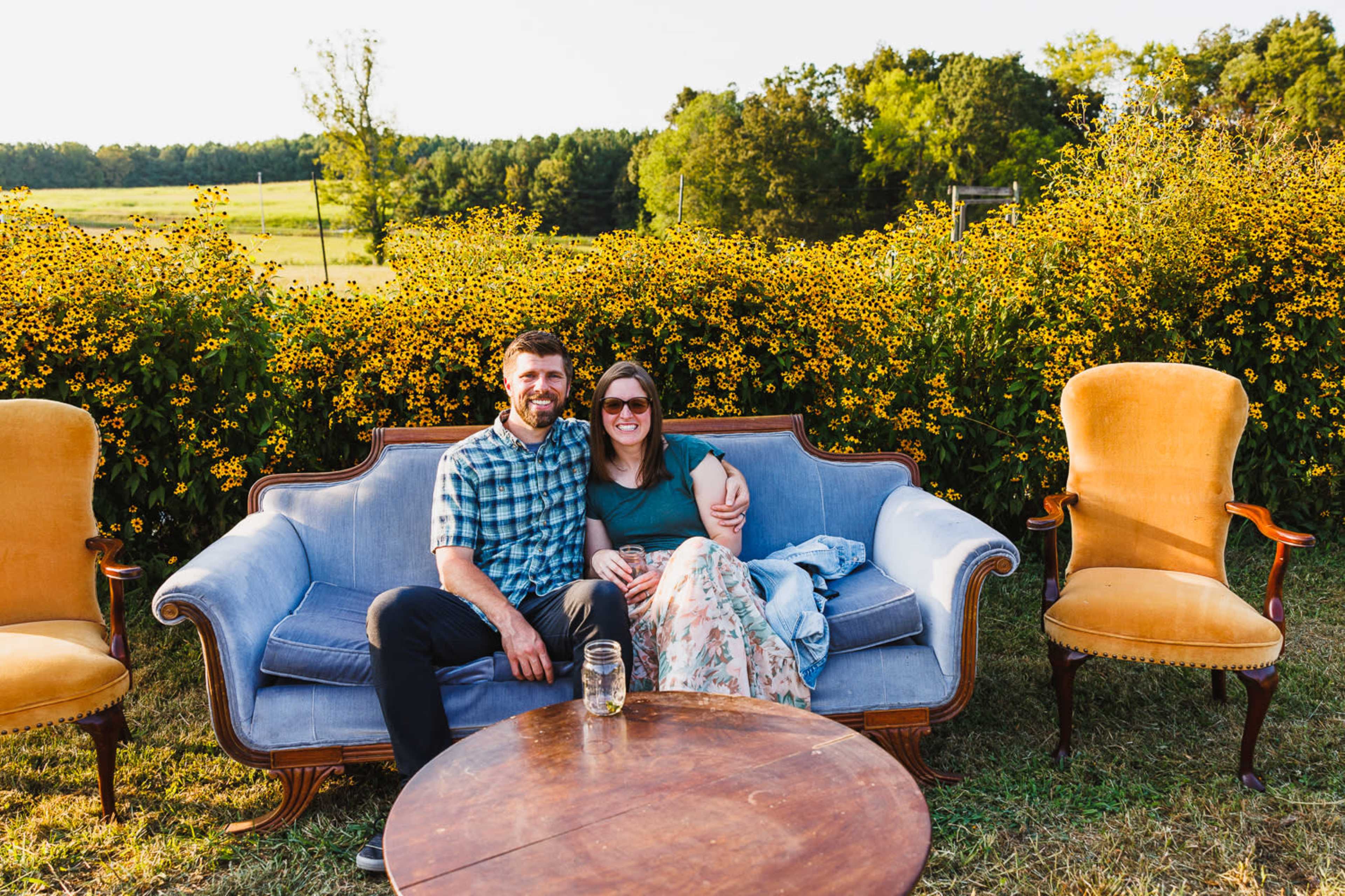 A couple sits on a vintage sofa surrounded by a field of yellow flowers, with a rustic table in front of them.