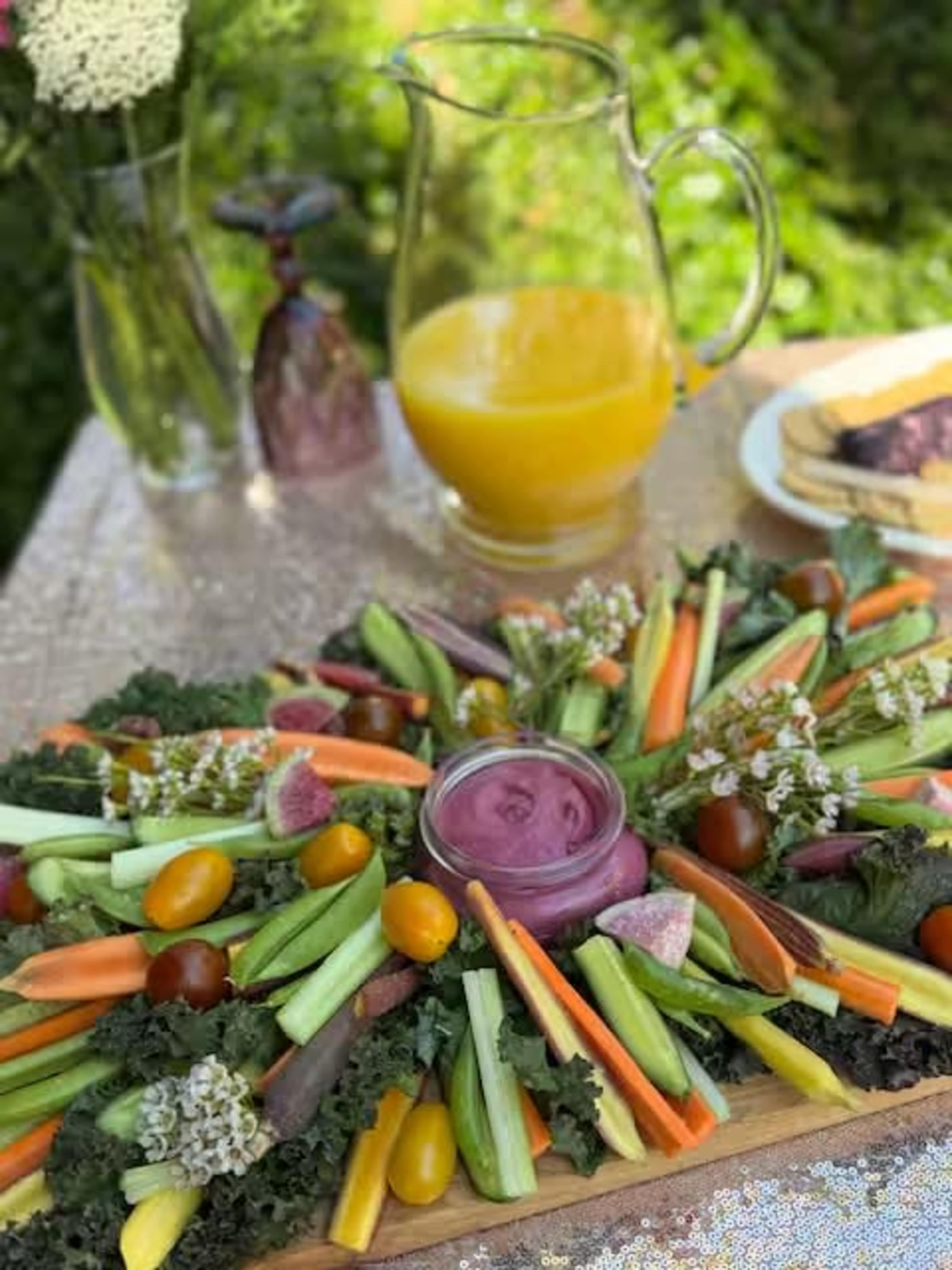 A vibrant vegetable platter arranged in a circular design with a dip in the center, accompanied by a pitcher of orange juice and some slices of bread.