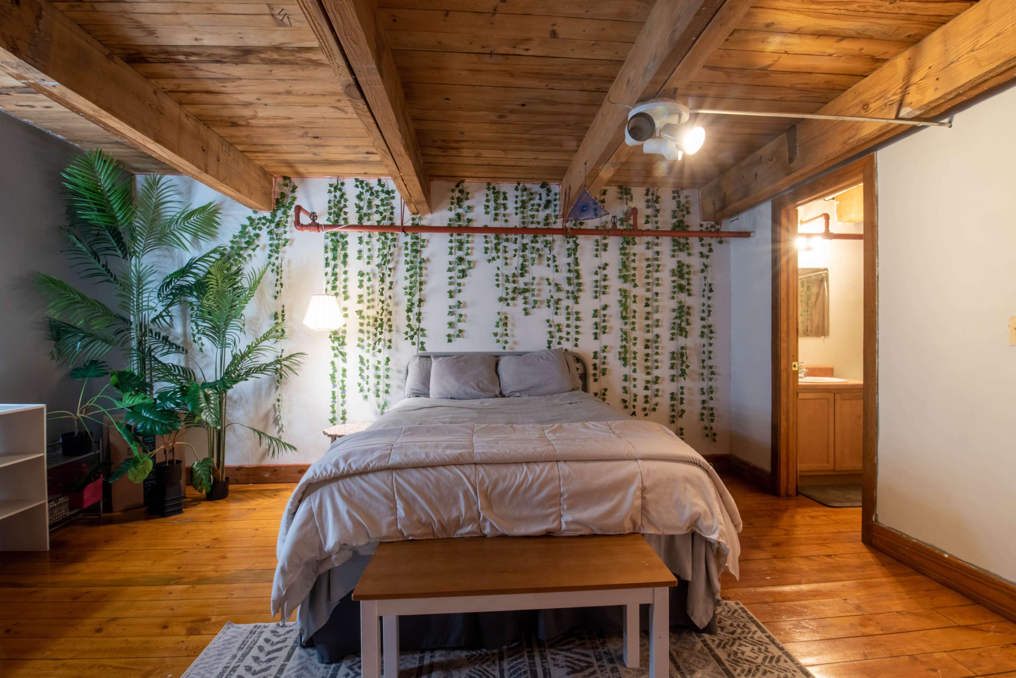 A bedroom with a wooden ceiling, a bed covered with a gray comforter, a bench at the foot of the bed, and potted plants against a wall adorned with green vines.