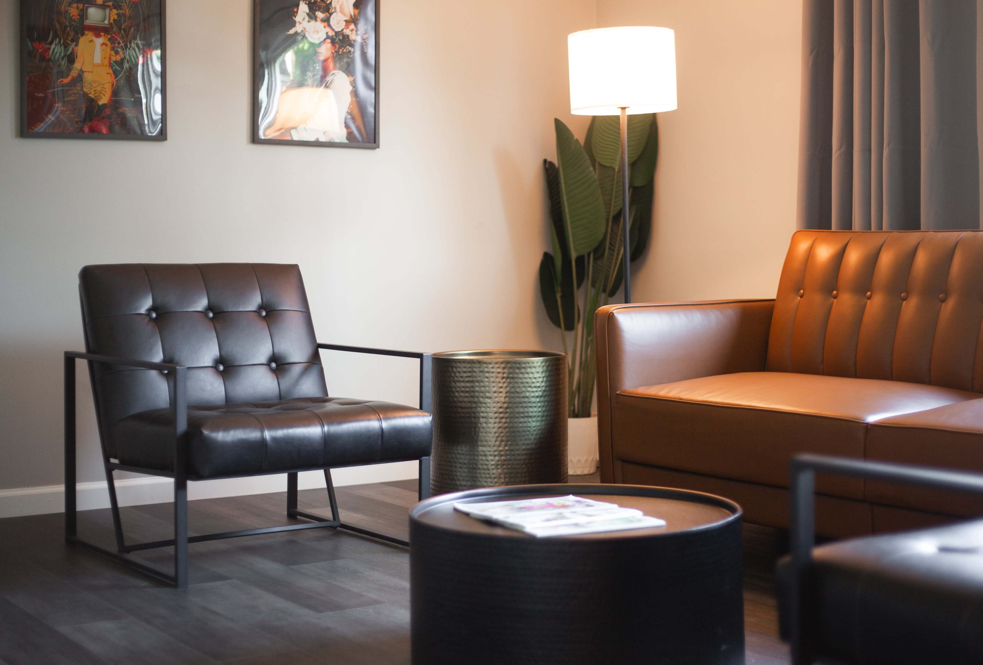 The scene features a modern living room with a leather sofa, a black accent chair, a round coffee table, and a standing lamp beside a potted plant.