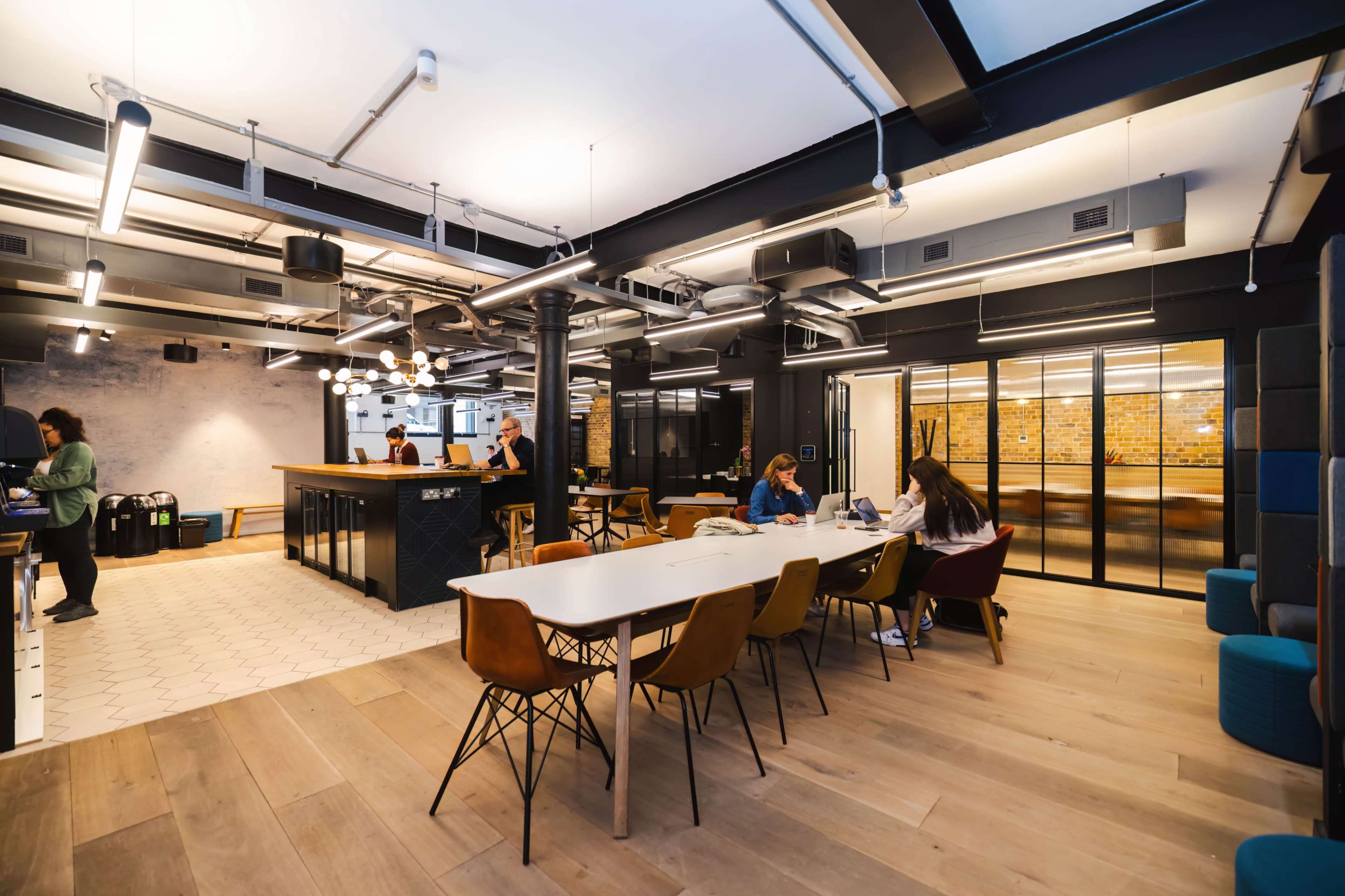 The image shows a modern office space with a communal dining area, workspace, and glass-walled meeting rooms, featuring people engaged in various activities.