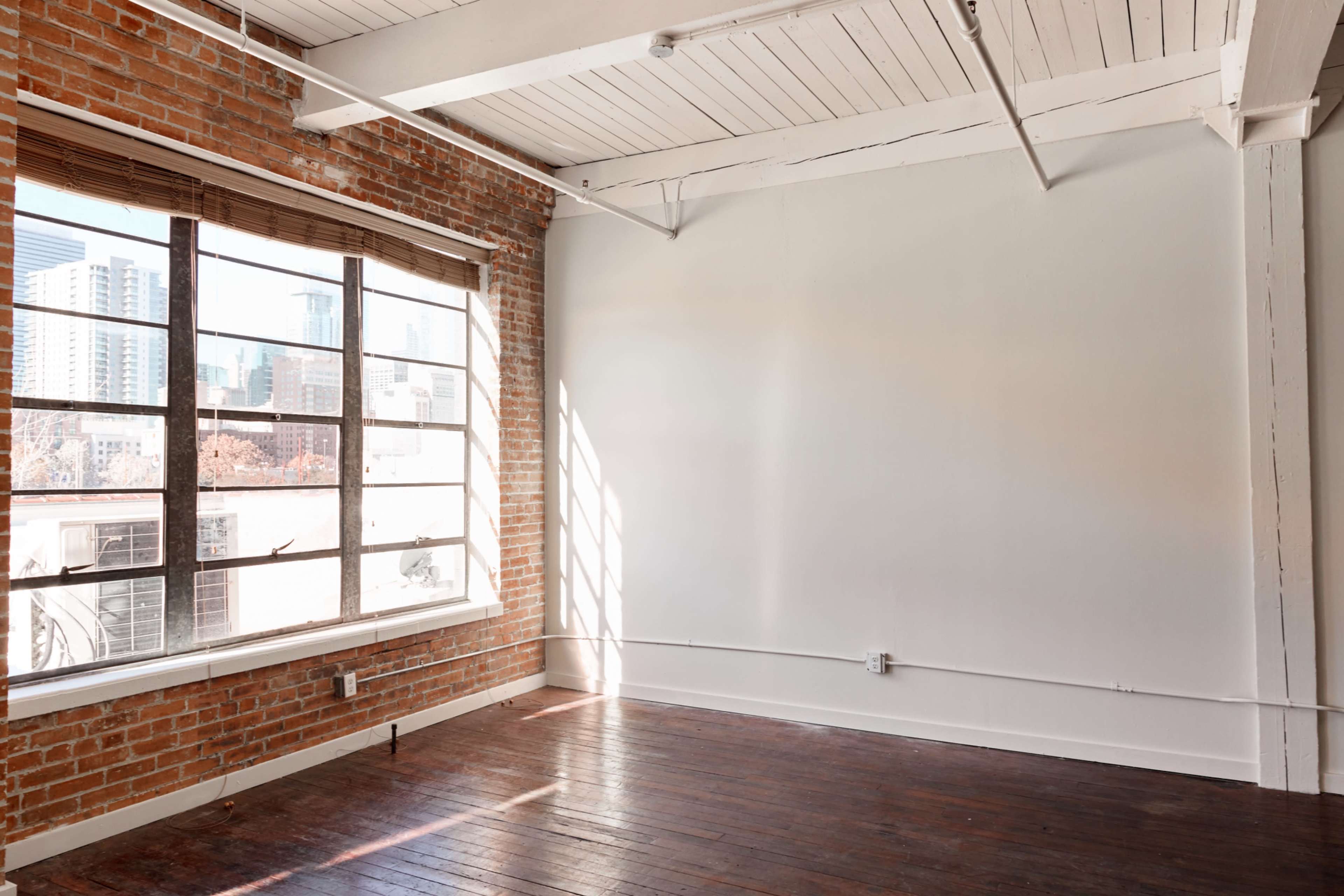 An empty room with exposed brick walls, a large window allowing natural light, and a wooden floor.