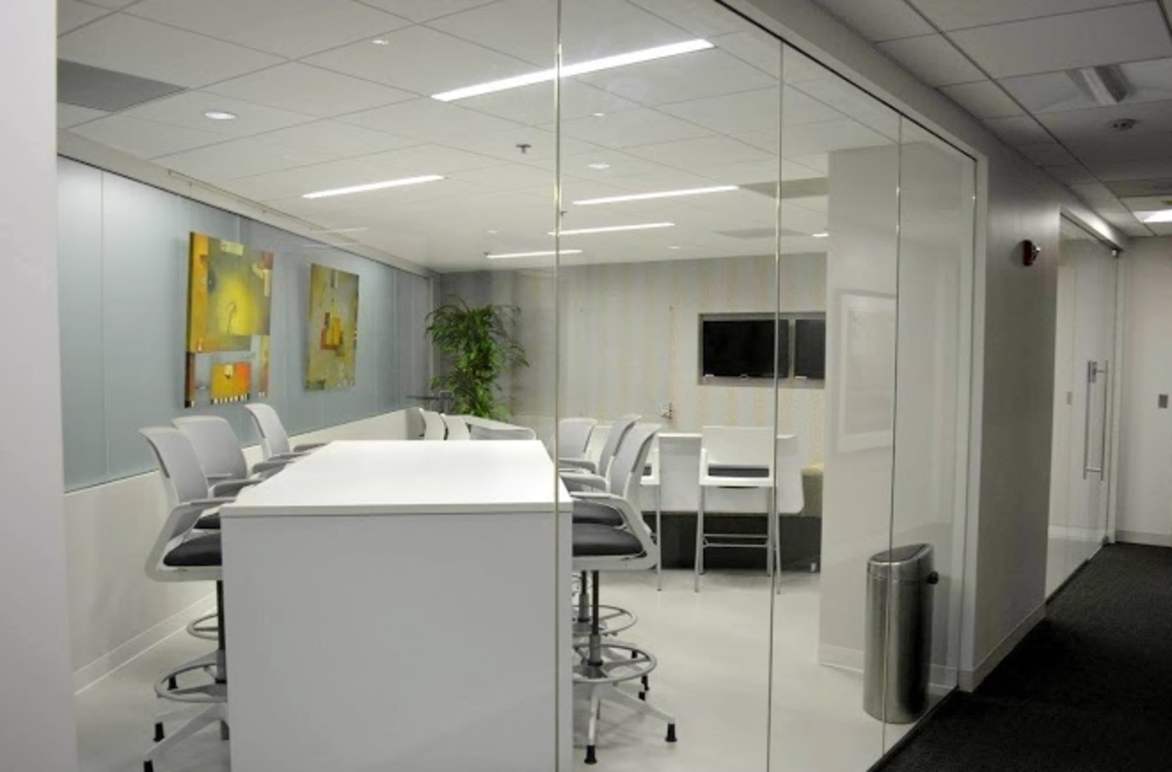 A modern office meeting room features a glass wall, white furniture, and wall art.