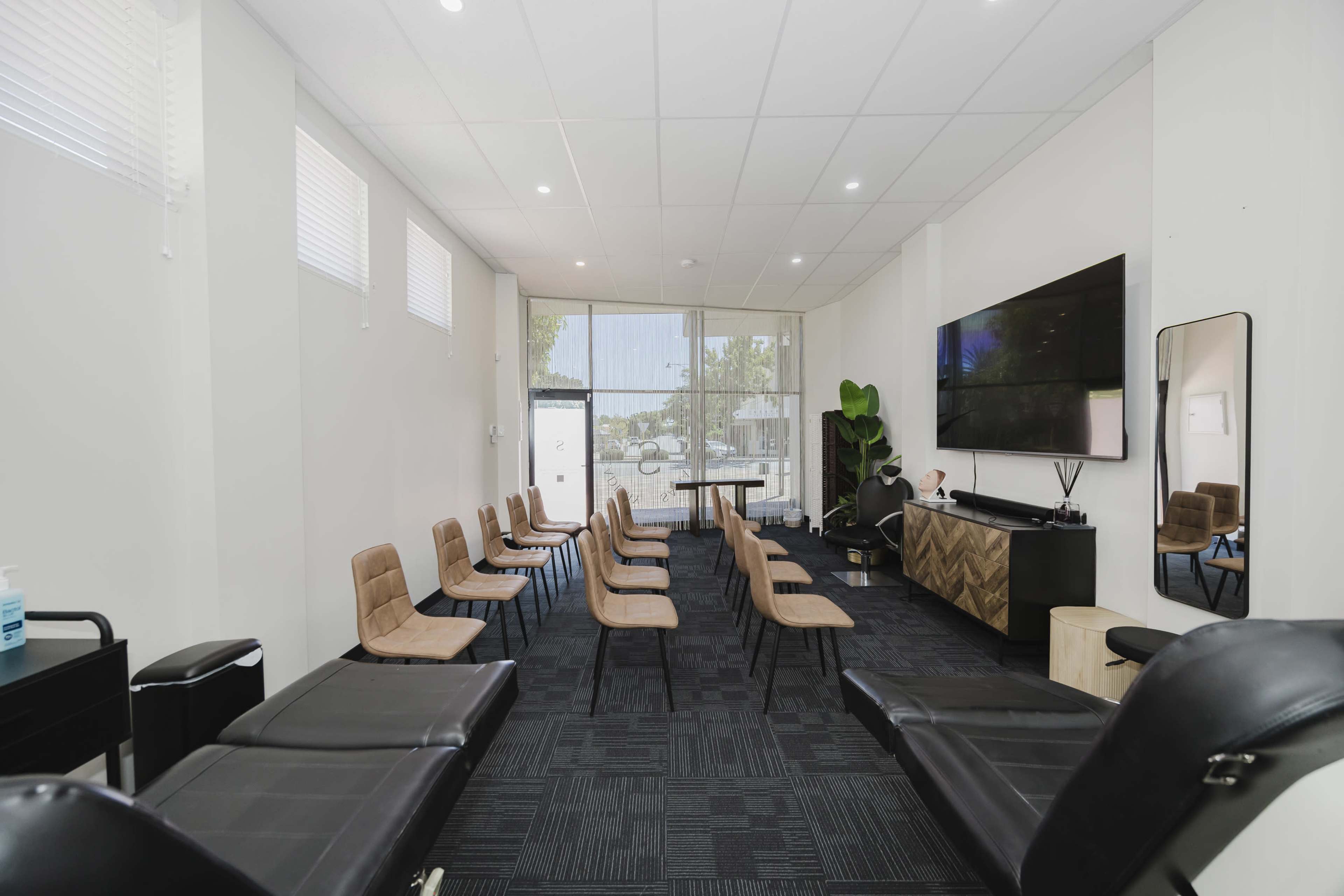 The image shows a spacious, modern waiting area with several rows of chairs, a large television, and a view of a street through large windows.