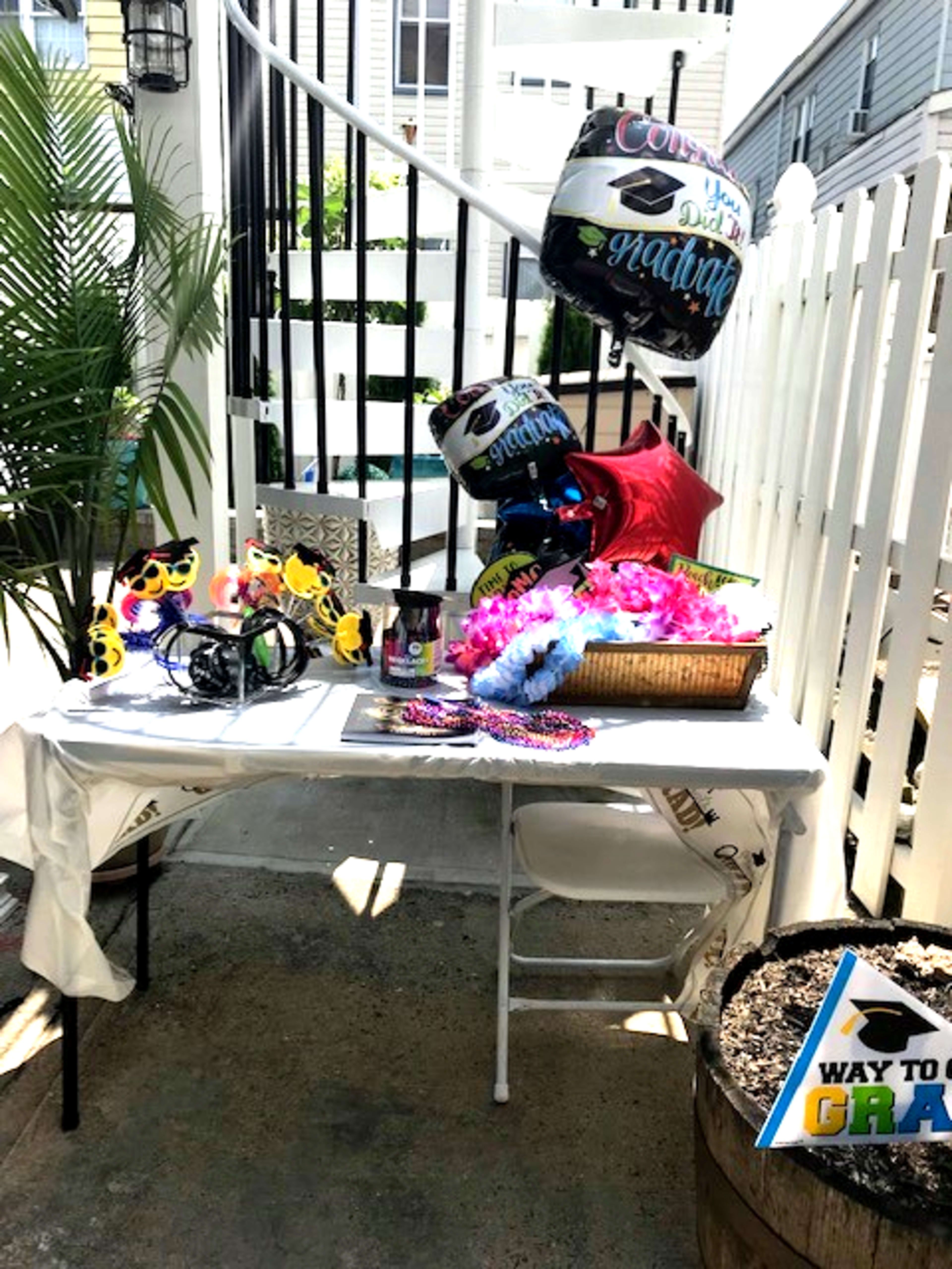 A table is set up with graduation-themed decorations, including balloons, hats, and colorful leis, beside a staircase and a white fence.