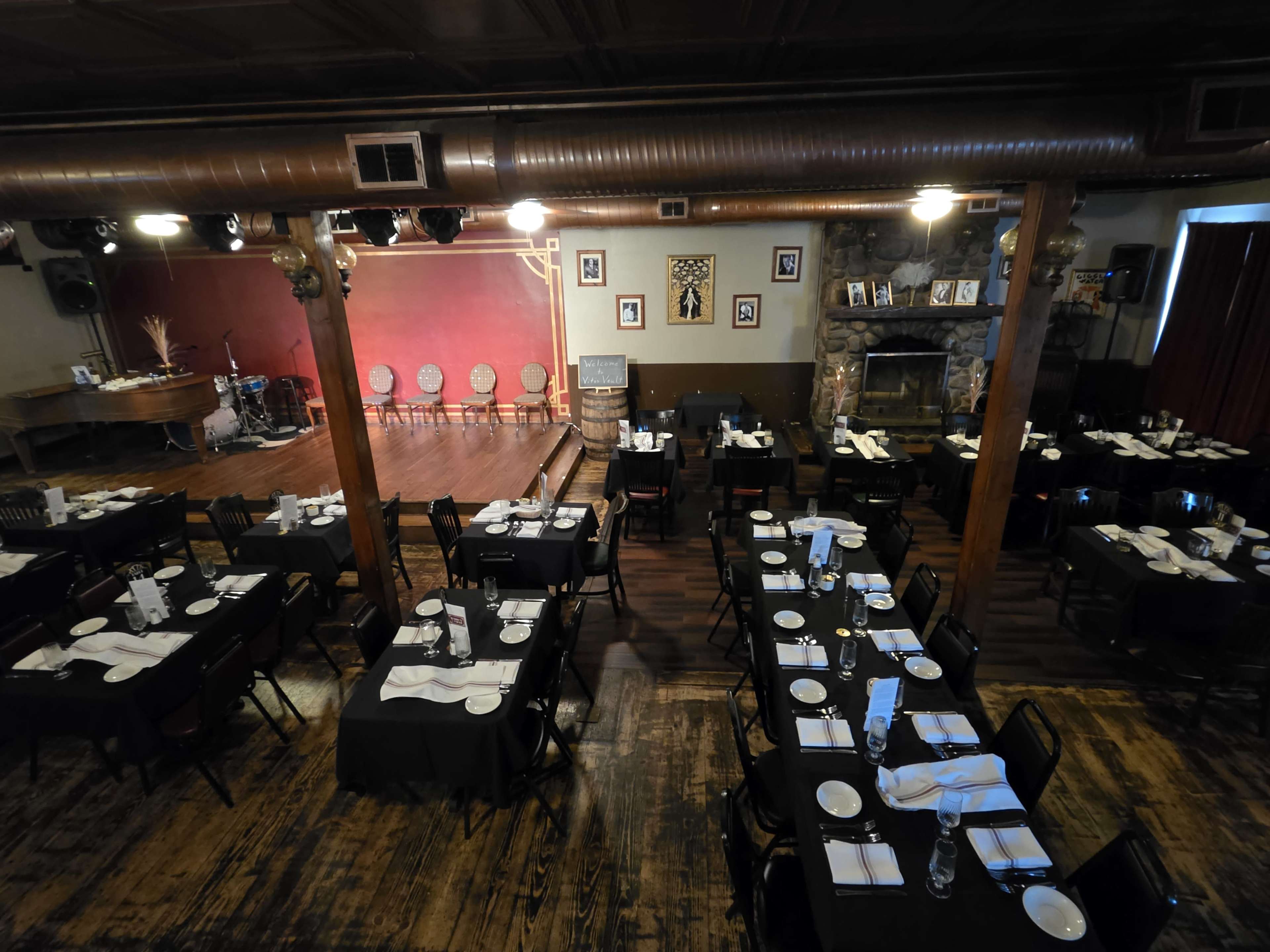 The image shows an elegant restaurant with black tables set for dining, arranged in rows facing a small stage, and featuring wooden beams and a stone fireplace.