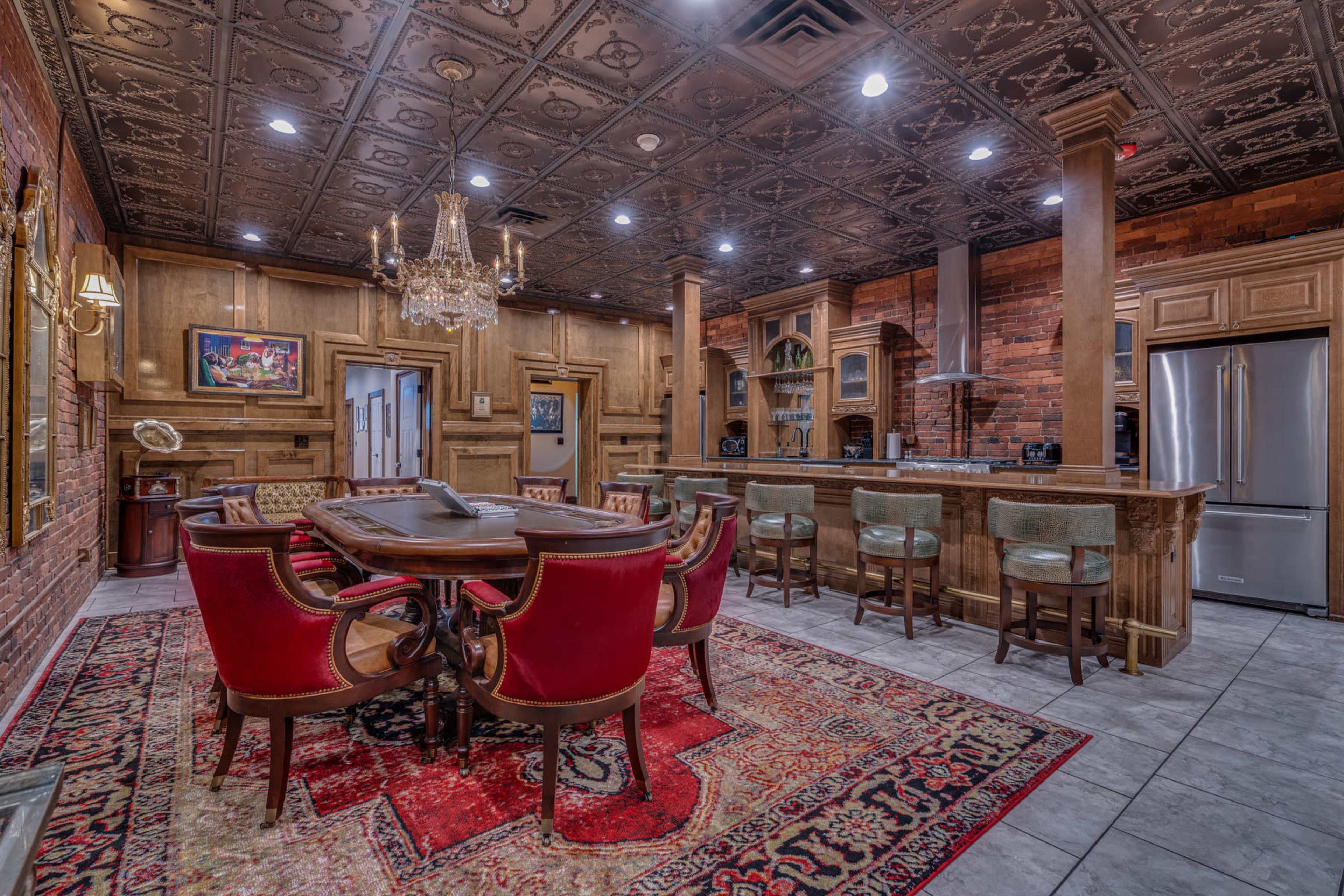 The image shows a spacious dining area with a wooden table surrounded by red upholstered chairs, a chandelier overhead, and a kitchen area featuring stainless steel appliances and wooden cabinetry.