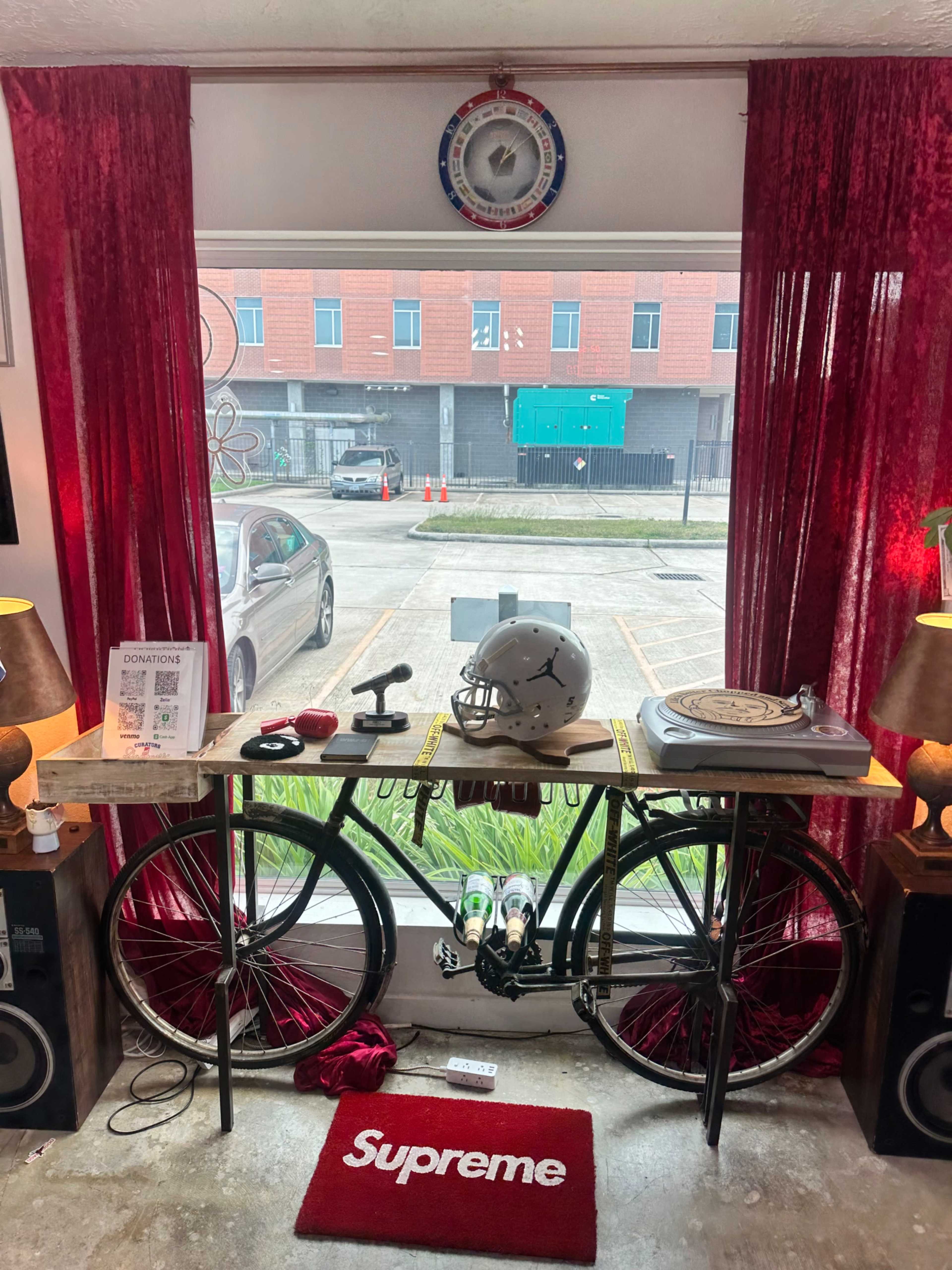 An indoor scene features a bicycle displayed under a table with a red carpet, two lamps, and a vintage clock, all framed by red curtains and a view of a city street outside.