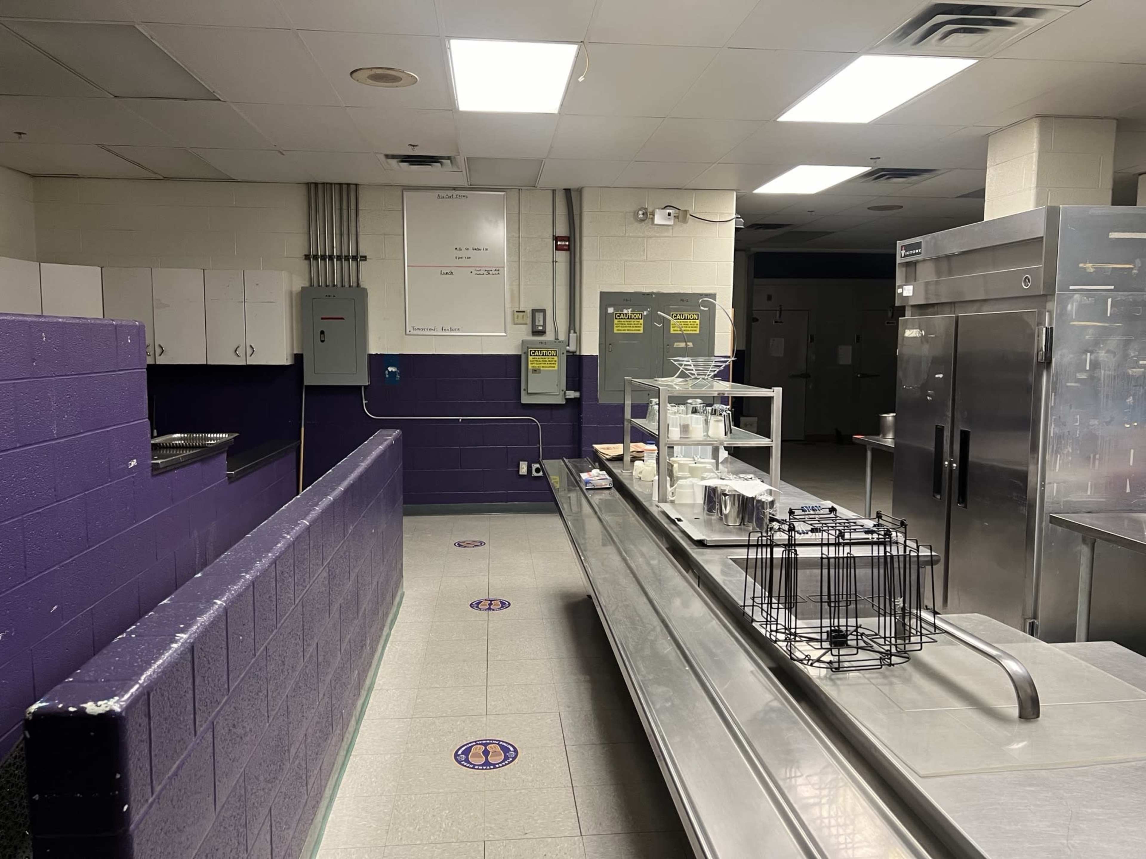 The image shows a kitchen area with purple and gray walls, metal countertops, and large refrigerator units, featuring a serving line and storage shelves.