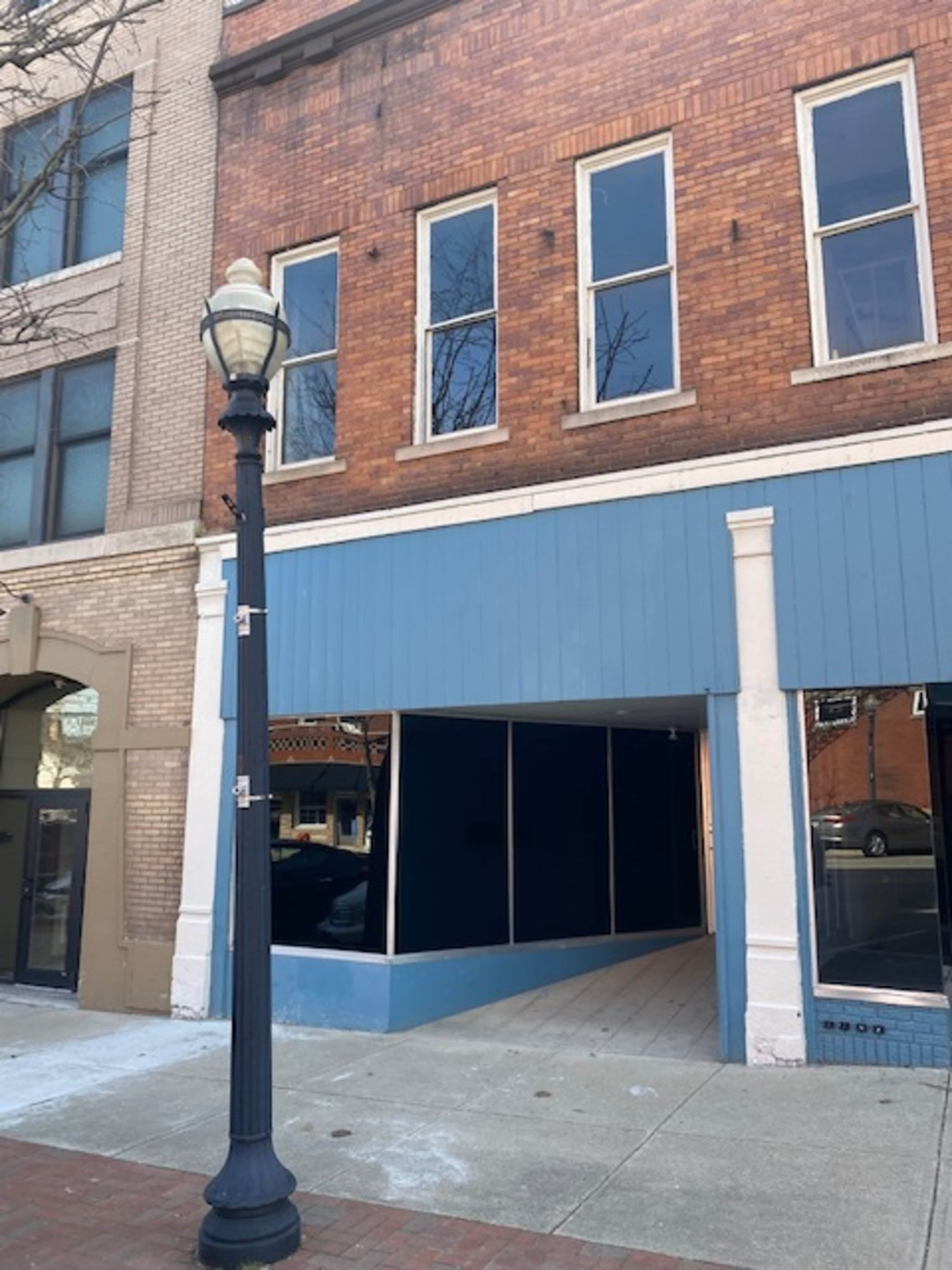 The image shows a vacant storefront with large glass windows and a blue facade, located next to a streetlamp on a city sidewalk.