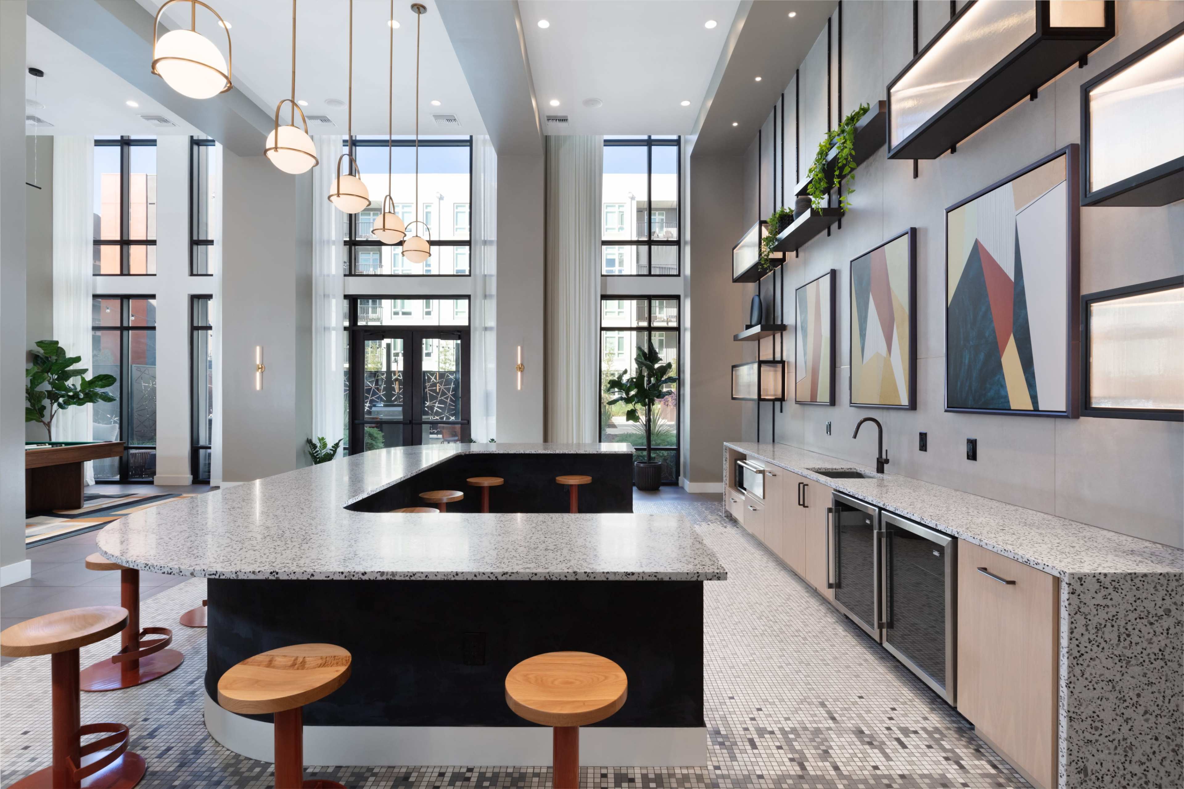 The image depicts a modern kitchen area featuring a large granite countertop with wooden stools, large windows allowing natural light, and geometric wall art.