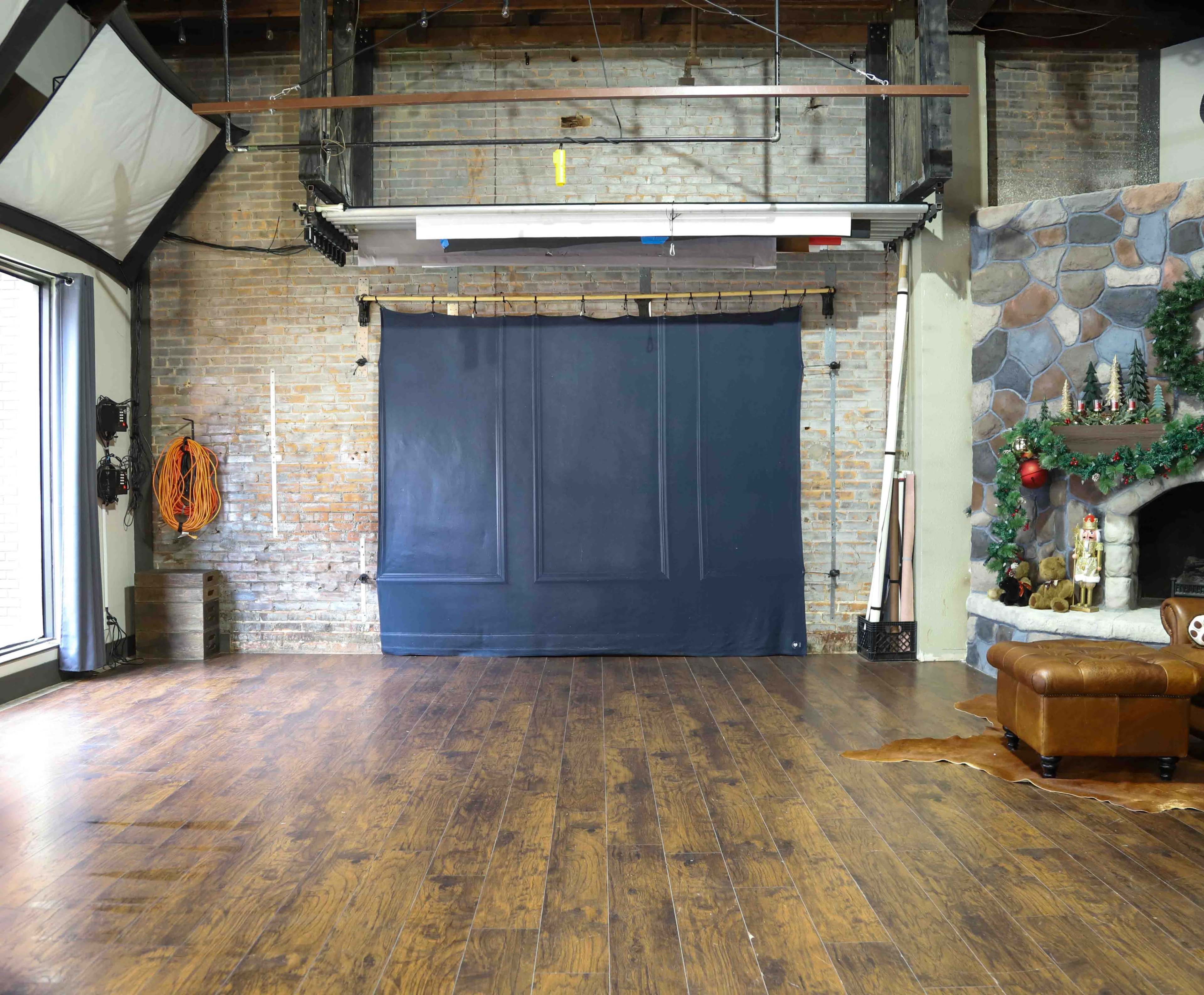 The scene features an empty studio space with a dark backdrop, wooden flooring, and a stone fireplace decorated with holiday ornaments.