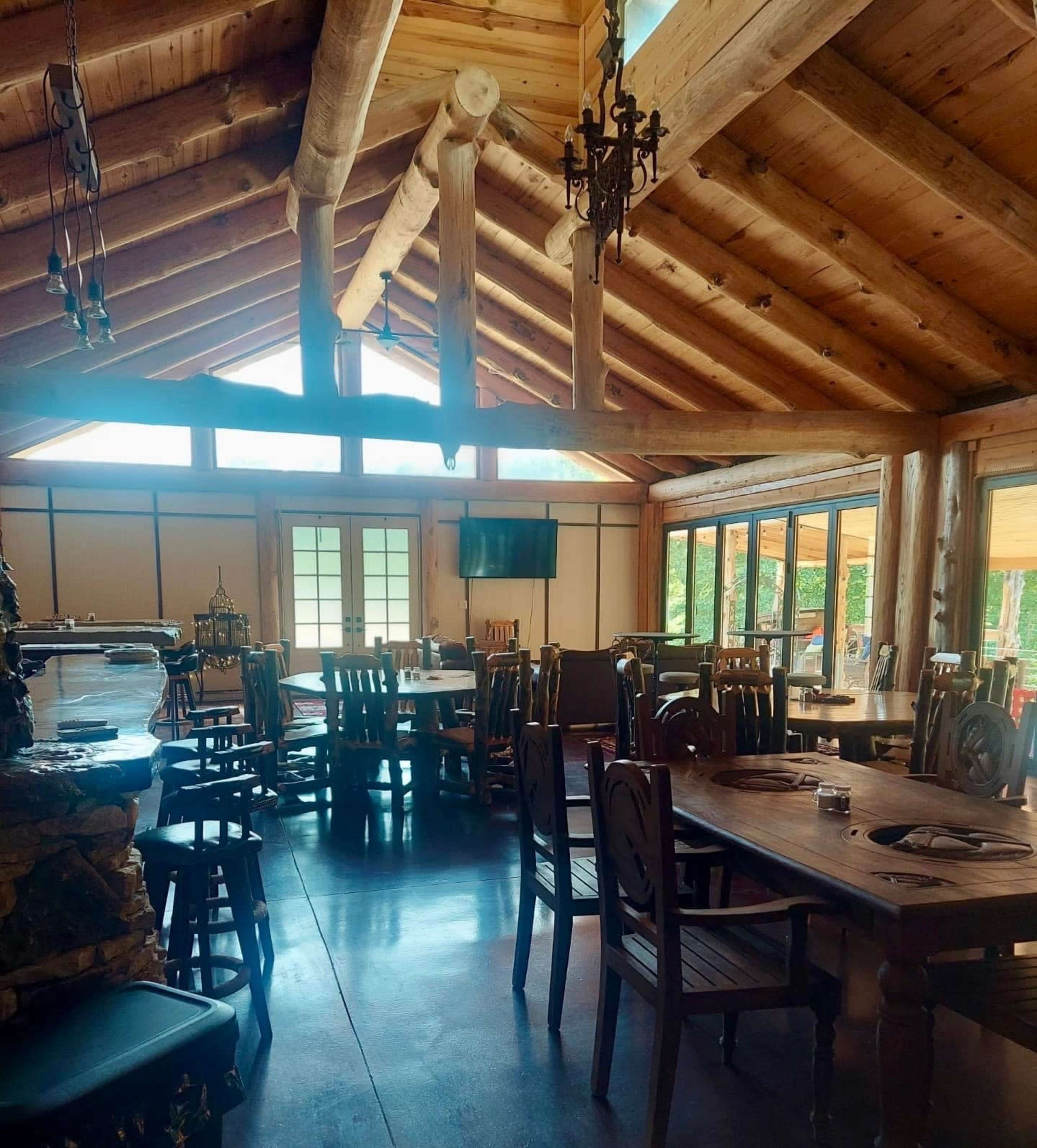The image shows a rustic dining area with wooden furniture and a high, beam ceiling, featuring large windows that allow natural light to enter.