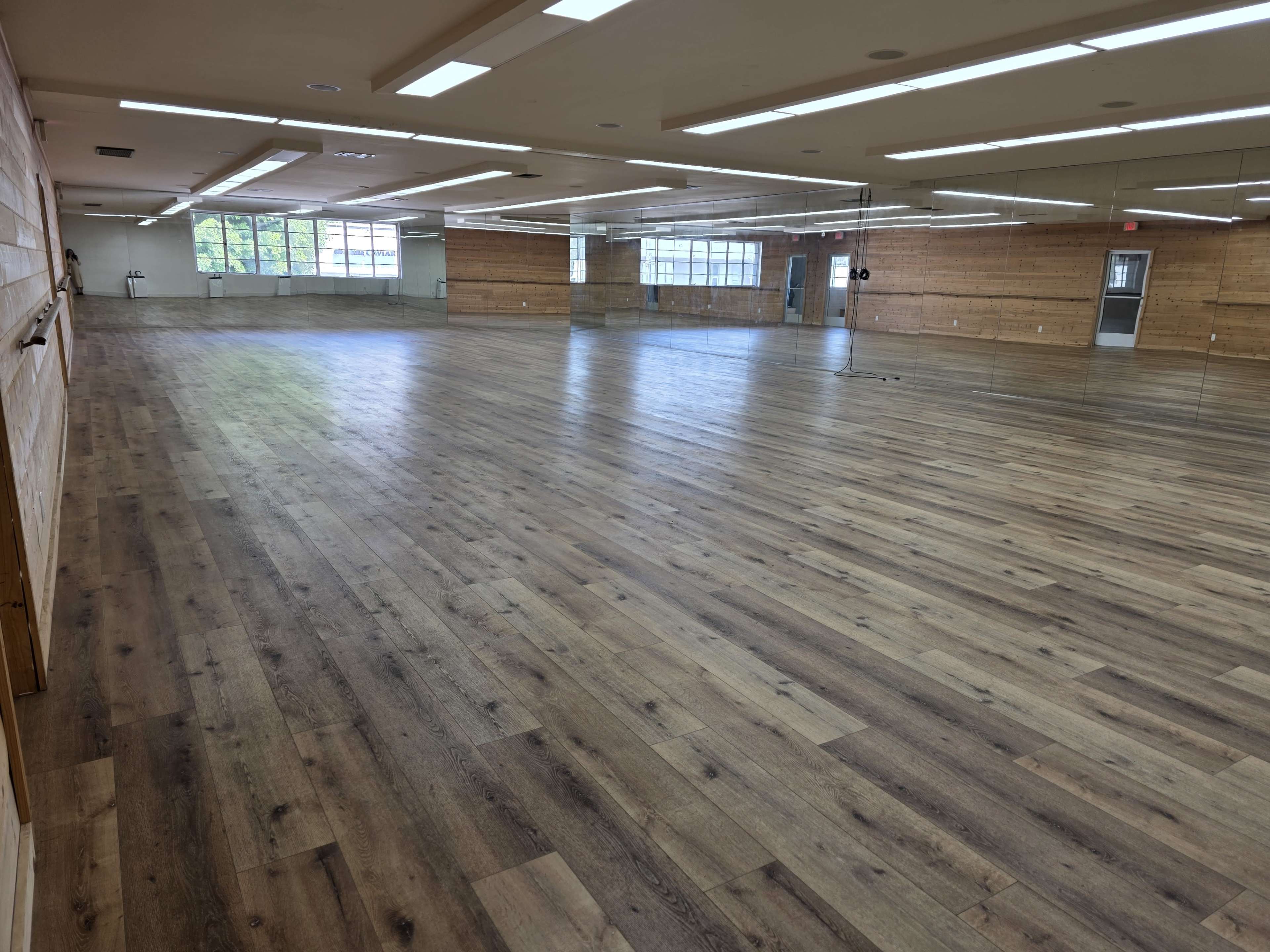 The image shows a spacious wooden-floored room with large windows and mirrors lining one wall.
