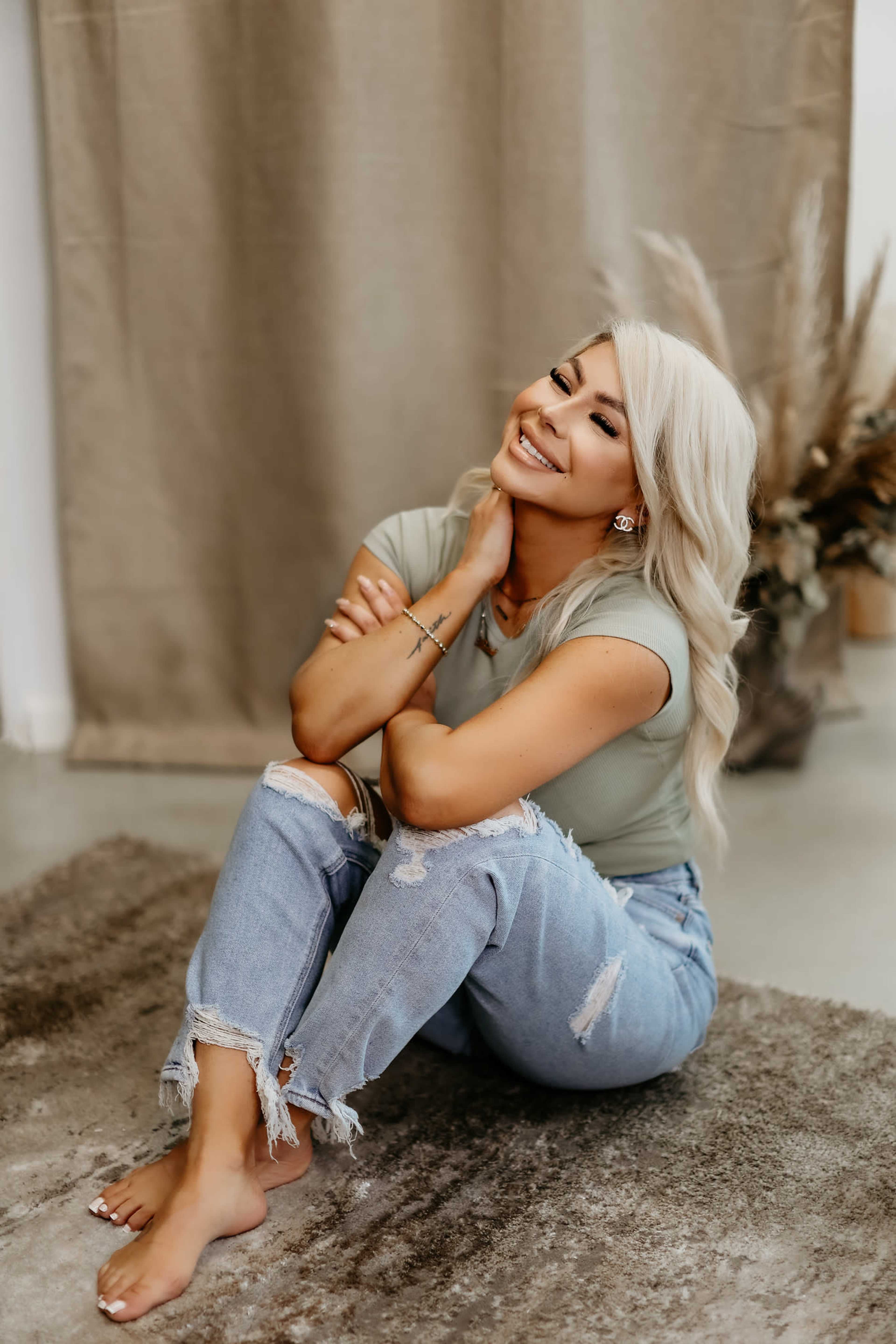 A woman with blonde hair sits on a rug, smiling while resting her chin on her hand, with a neutral backdrop and a decorative plant arrangement nearby.